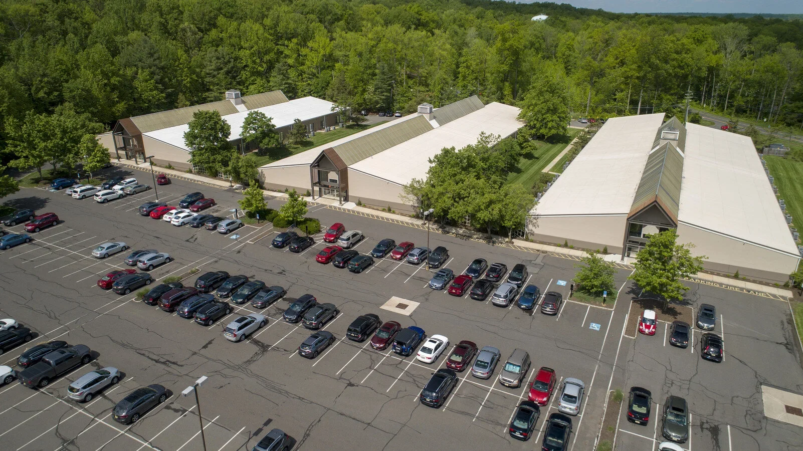 Aerial view of the Princeton Professional Park parking lot with numerous parked cars, surrounded by green trees, with a white building in the background.