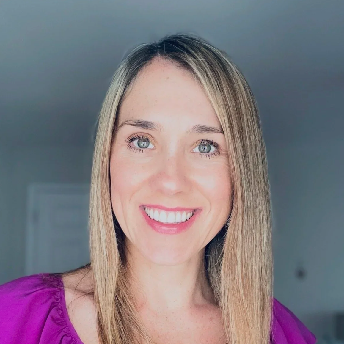 Close-up of a woman with long blonde hair, blue eyes, and a bright smile, wearing a purple top, indoors with a neutral background.