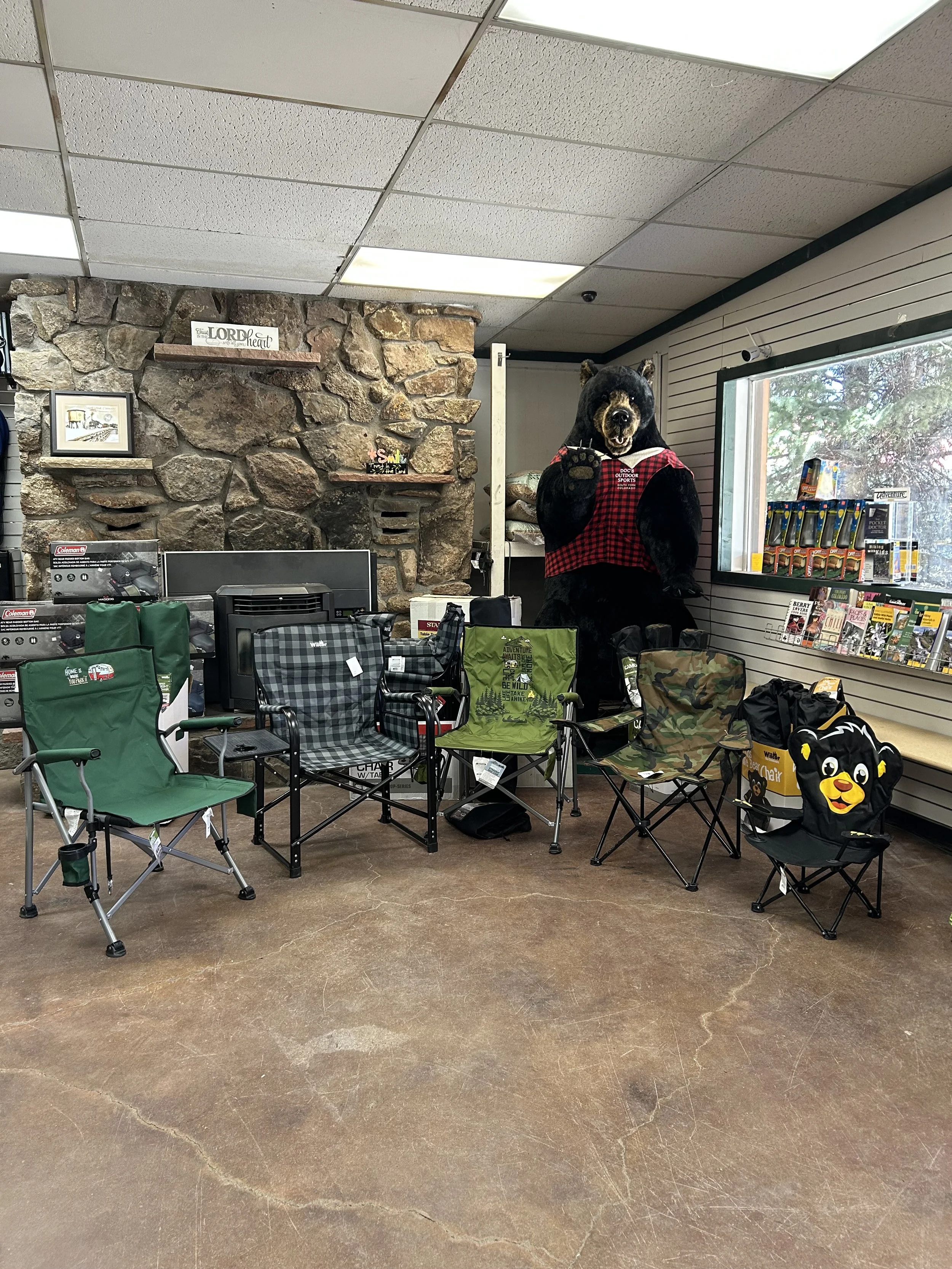 Store display with a large bear mascot dressed in red plaid shirt, sitting behind a counter with various camping chairs and outdoor gear in front, stone fireplace in the background, and magazines on the side.