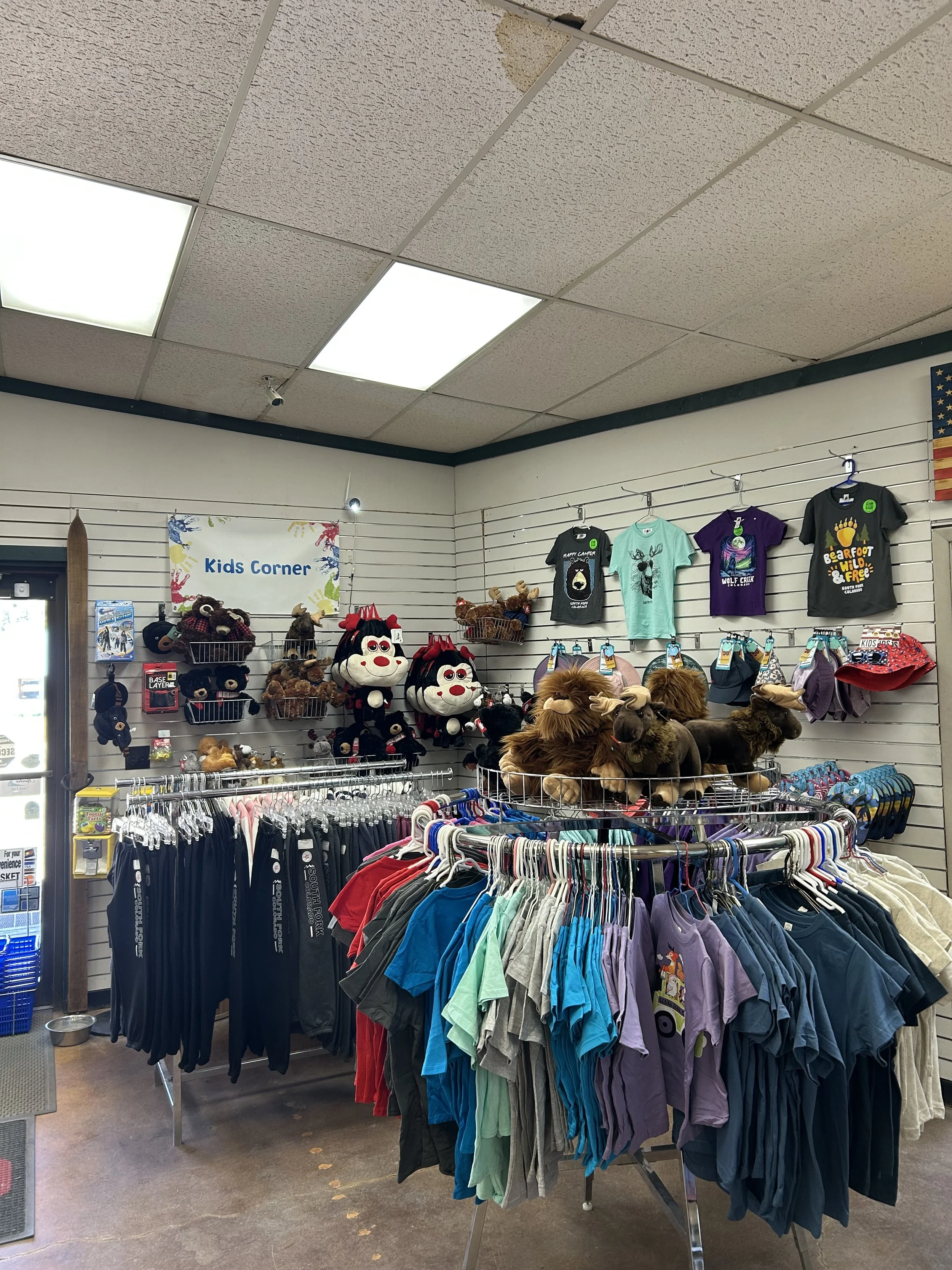 Display of kids' clothing and stuffed toys in a retail store, with a sign that reads 'Kids Corner.' T-shirts with various designs hanging on the wall and racks with colorful shirts.