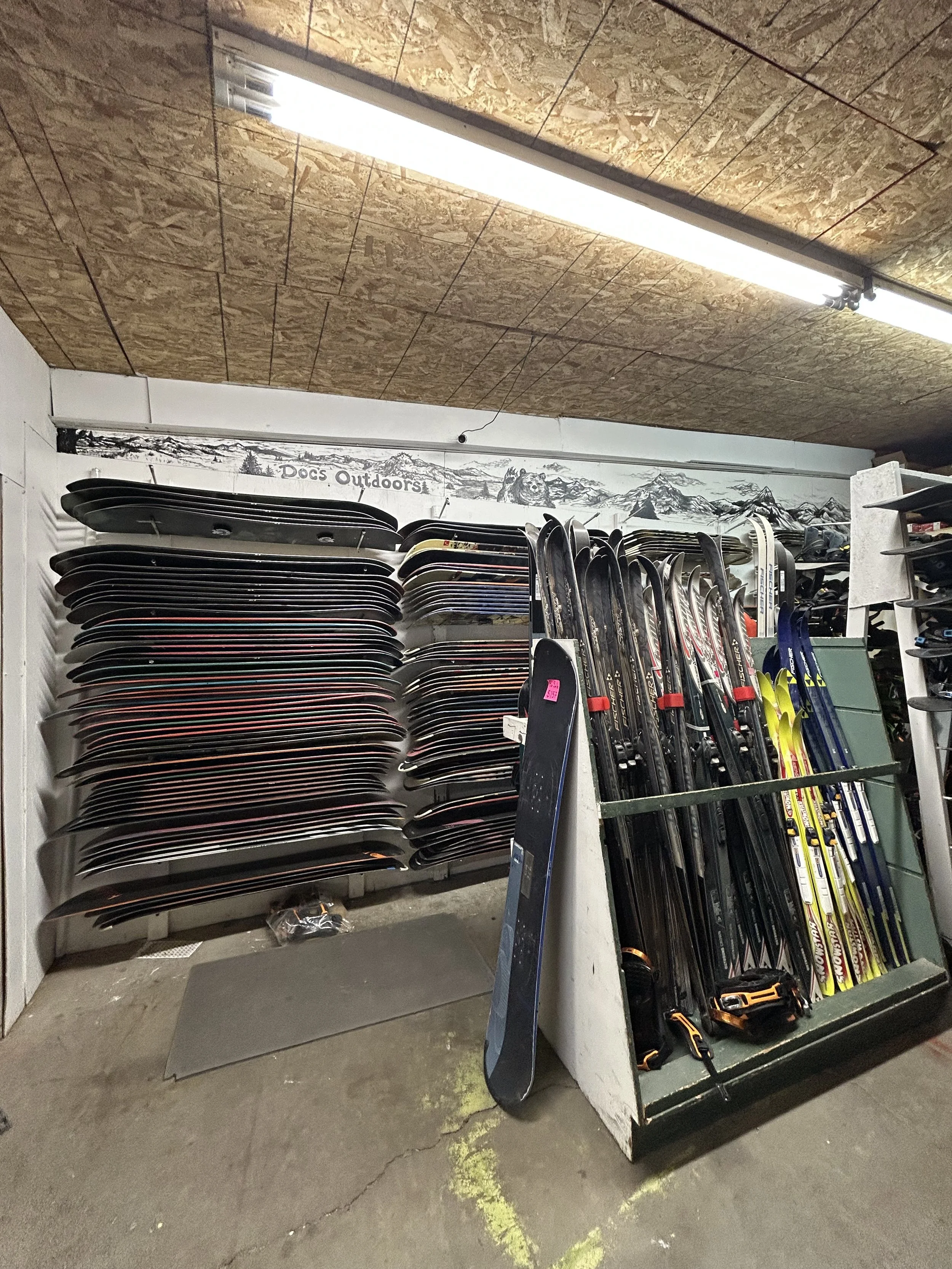 Wall of snowboards and skis on display in a sports store, with a mountain scene mural on the wall and fluorescent lighting overhead.