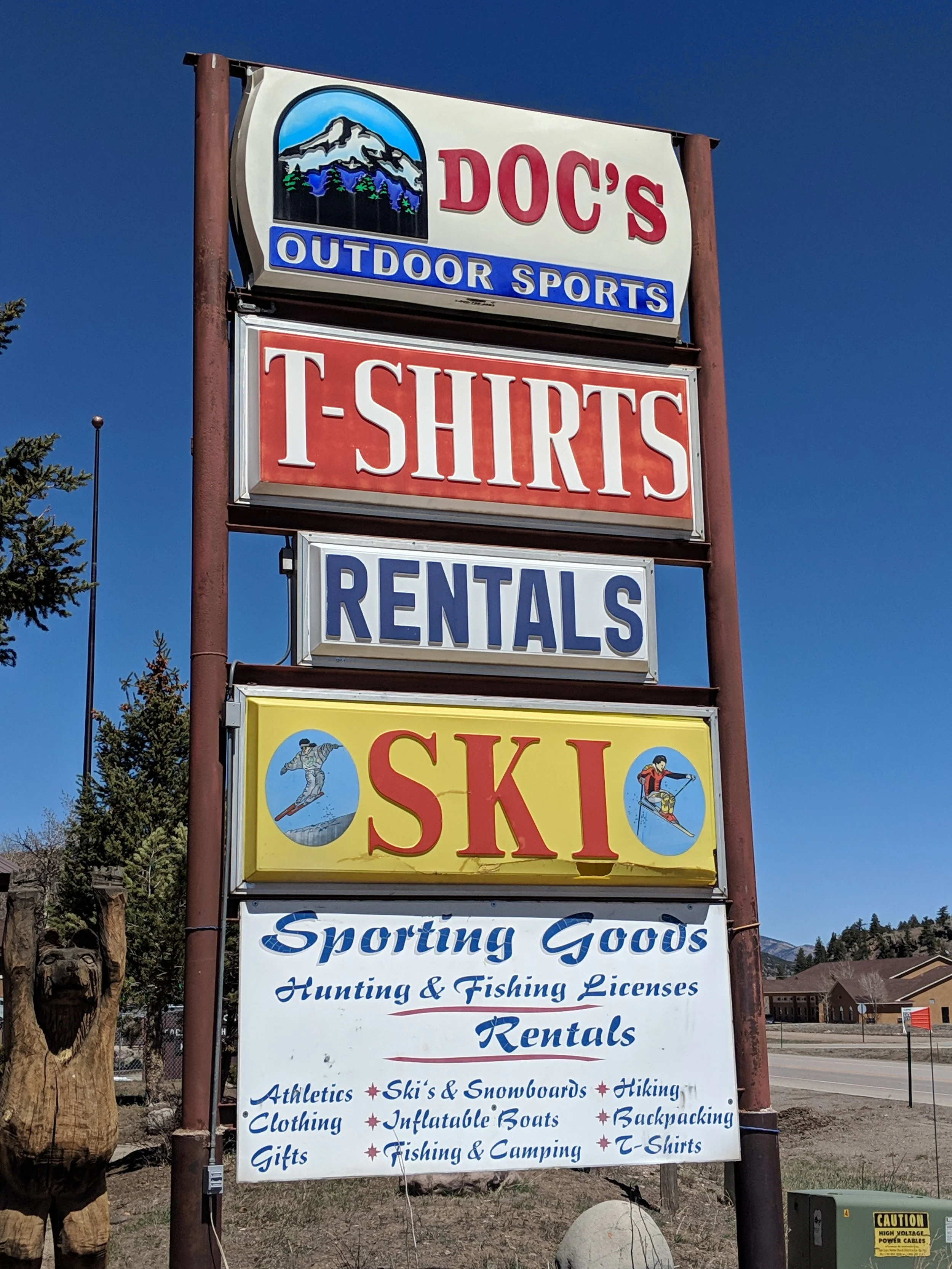 A tall outdoor sign with multiple sections. The top section has a mountain and forest logo with the text 'DOC'S OUTDOOR SPORTS'. The middle section has a red background with white text that reads 'T-SHIRTS'. The bottom section has white background with blue text advertising various outdoor activities and products, such as sporting goods, hunting and fishing licenses, rentals, and more.