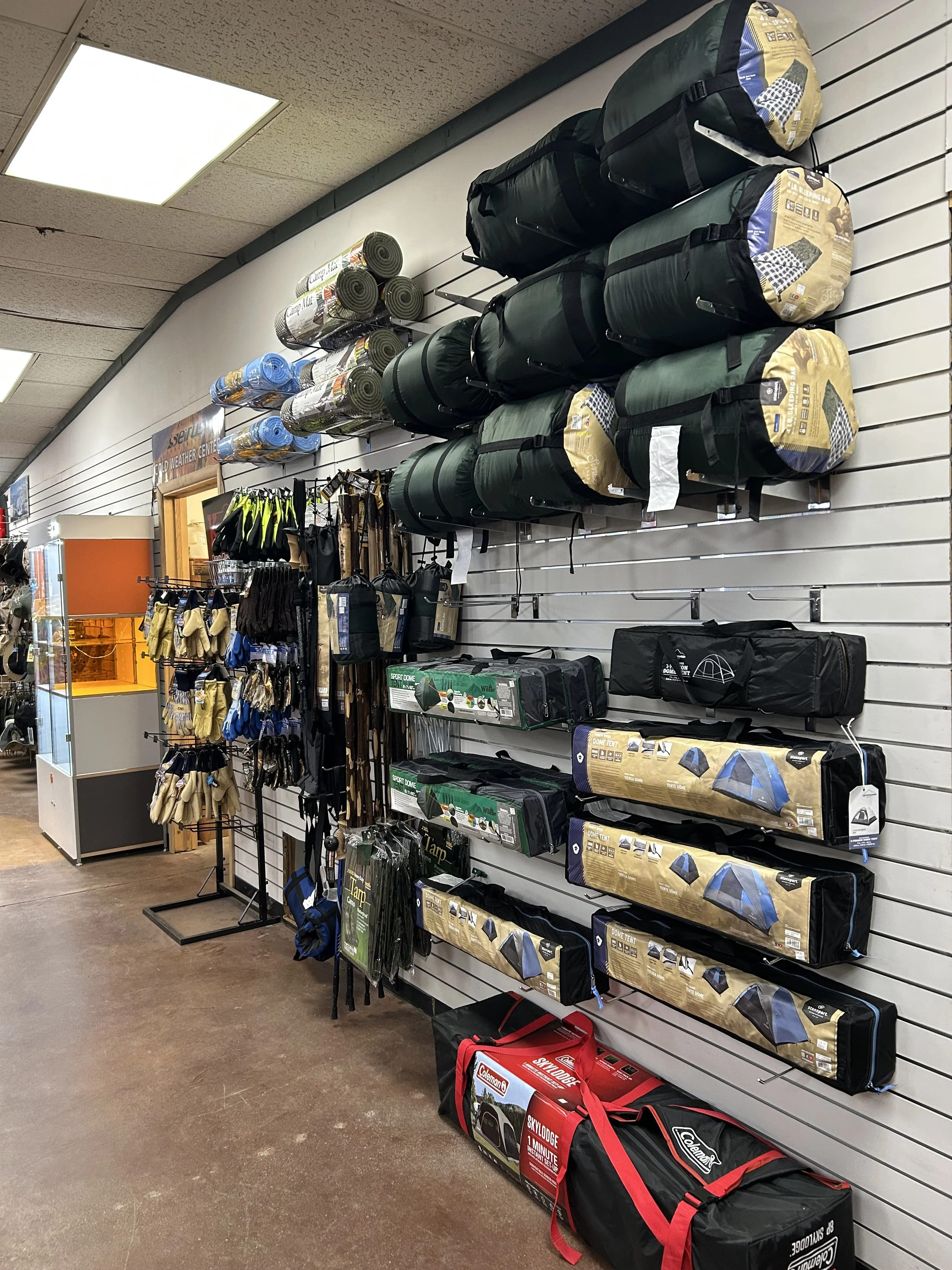 Store display with outdoor gear including camping tents, sleeping bags, and camping chairs on a slatwall, with gloves and other camping accessories hanging below.