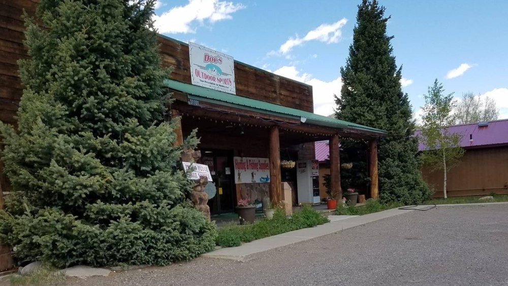 Wooden building with a green roof, signs for outdoor sports and supplies, large evergreen trees, and a partly cloudy sky.