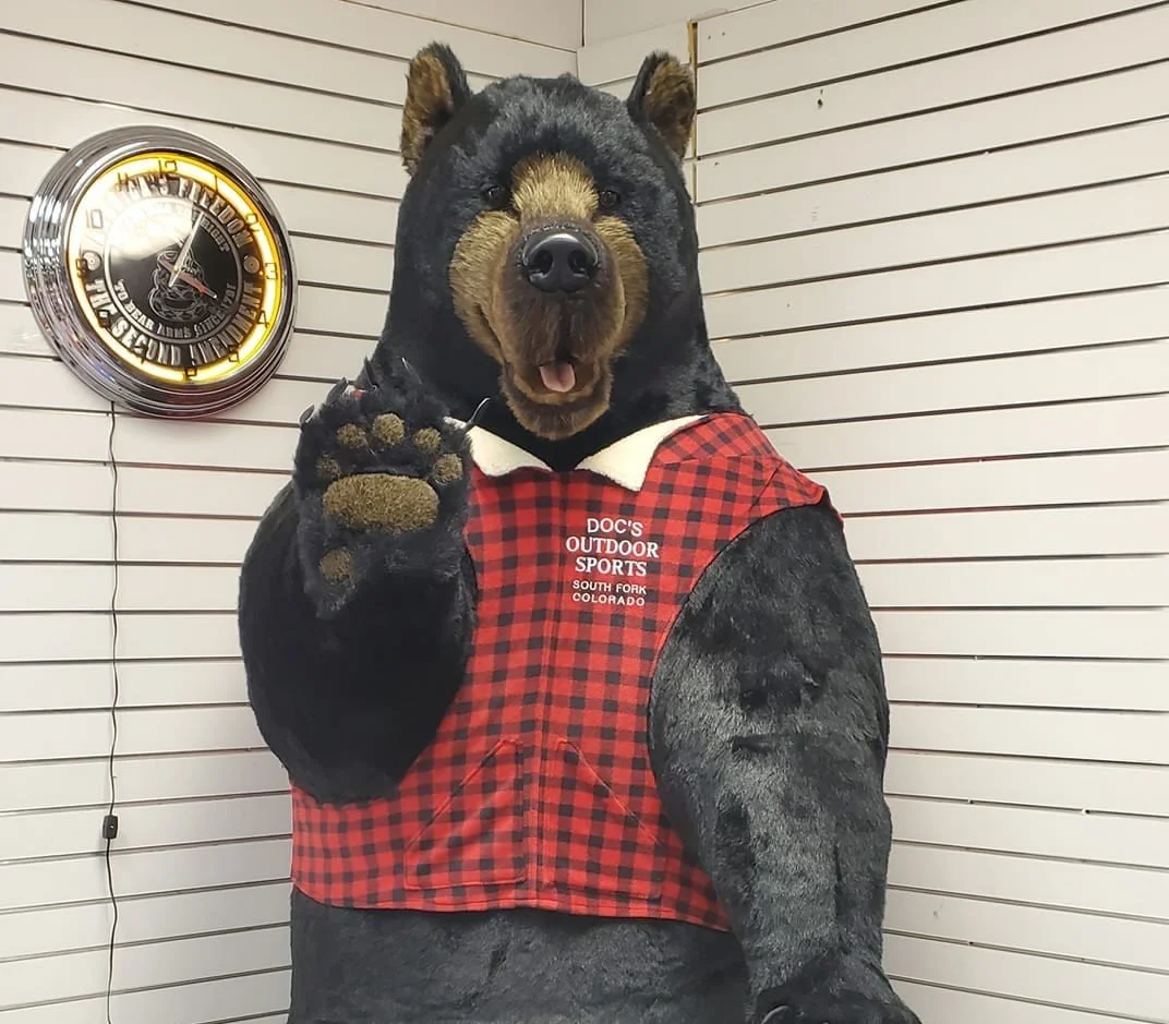 A person in a bear costume with a bear head and paws, wearing a red and black checkered vest that says 'Doc's Outdoor Sports South Fork Colorado,' is standing against a wooden wall with a clock on it.