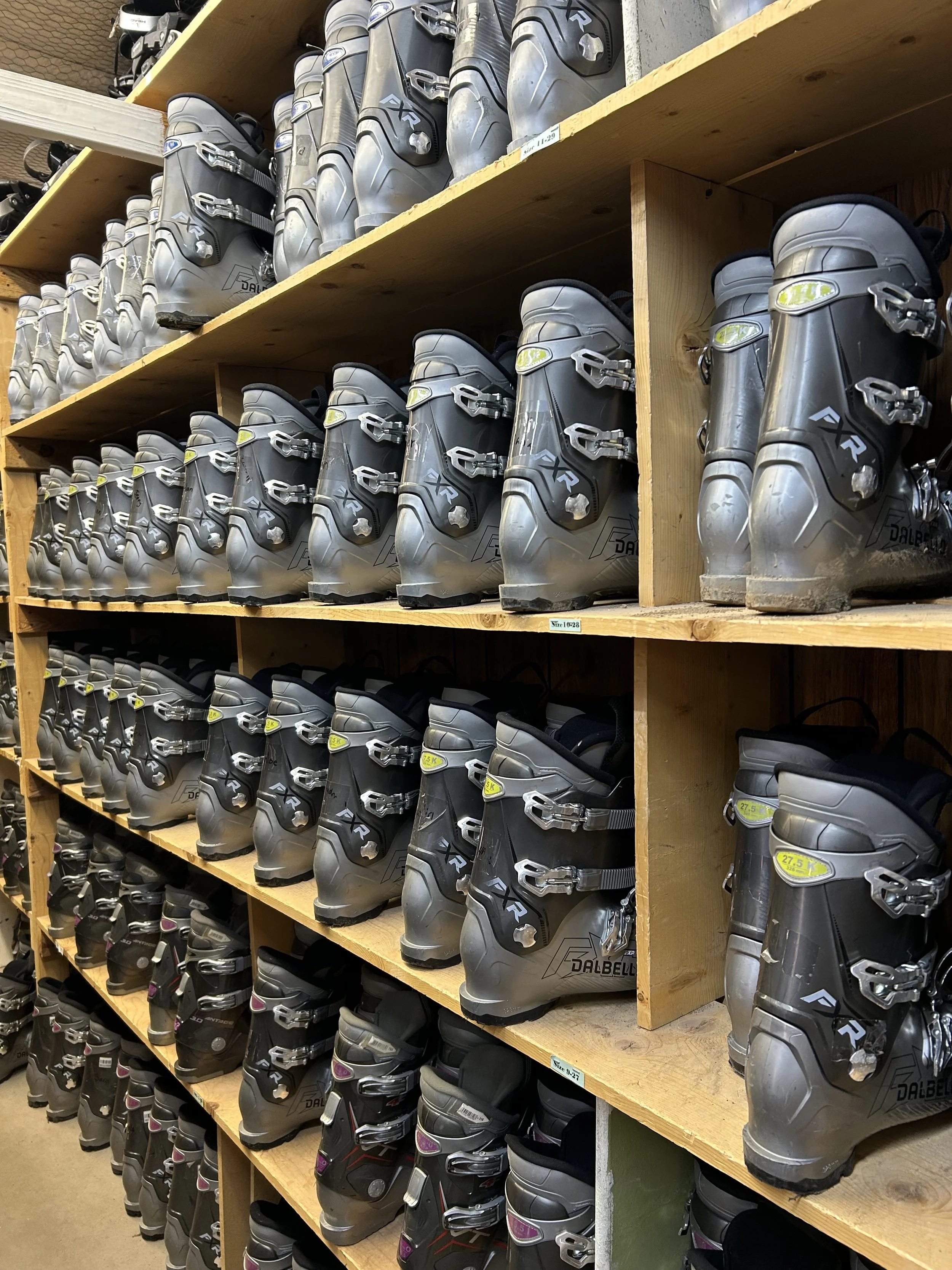 A store shelf filled with multiple pairs of ski or snowboard boots, arranged in rows and stacked on wooden shelves.