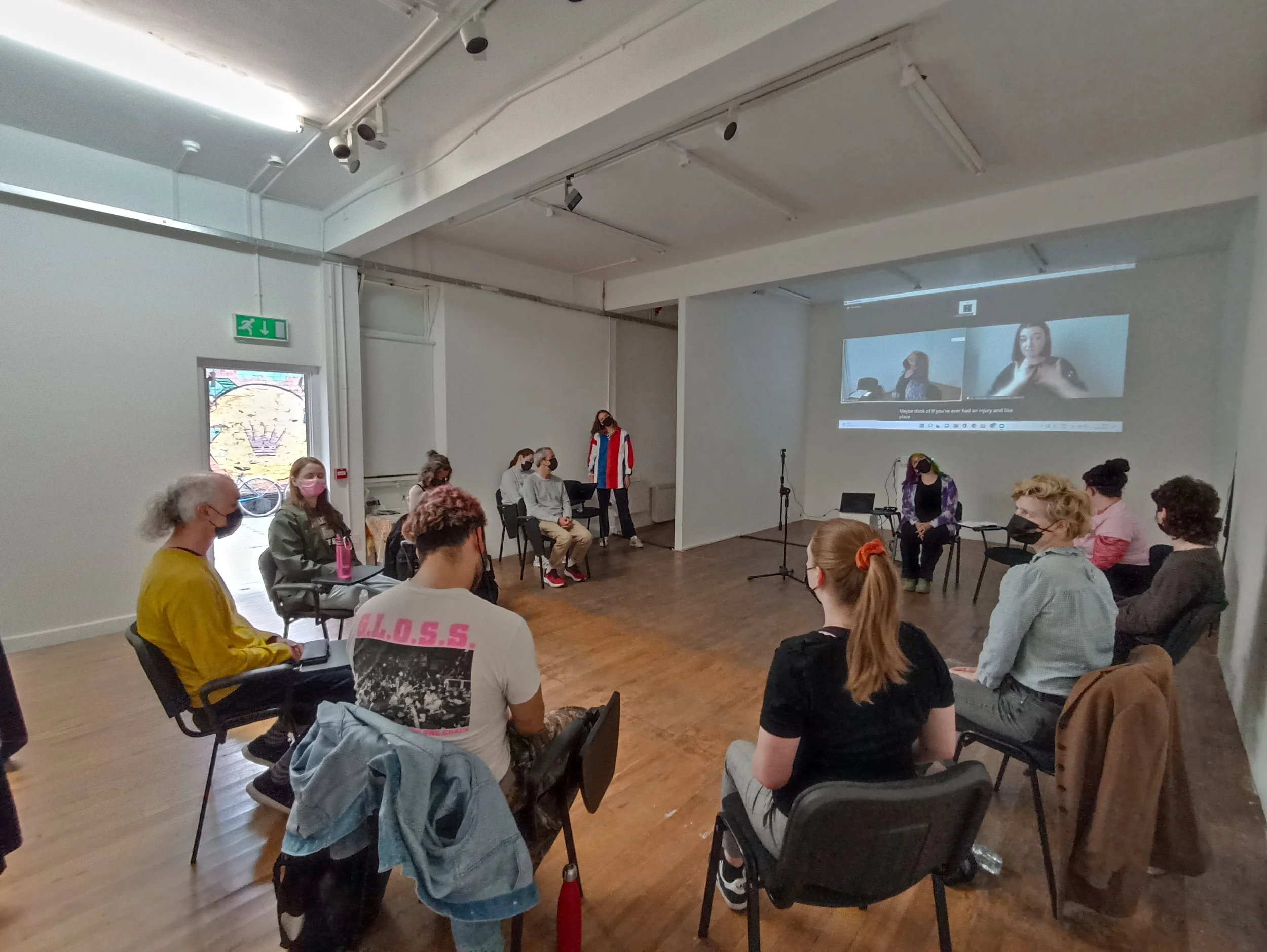 People seated in a well-lit room attending a presentation or discussion, with two people on the stage, one talking and the other sitting, and a large screen displaying a video call in the background.