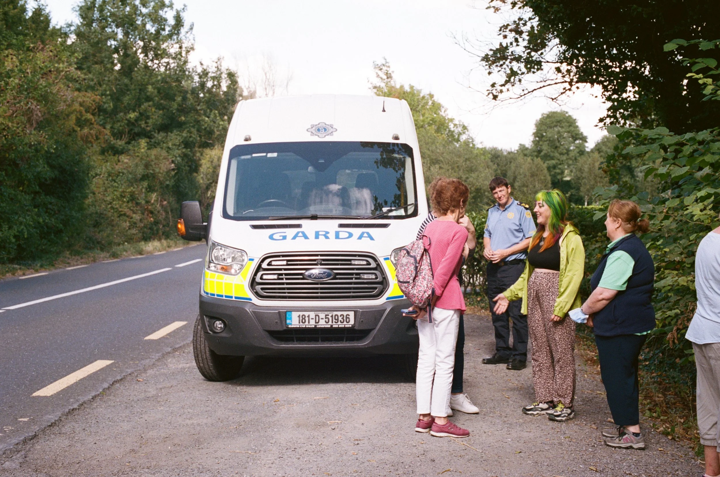 A group of five people standing on the side of a road in front of a Garda police van. The people are engaged in conversation, and the scene is set in a wooded area with trees and greenery.