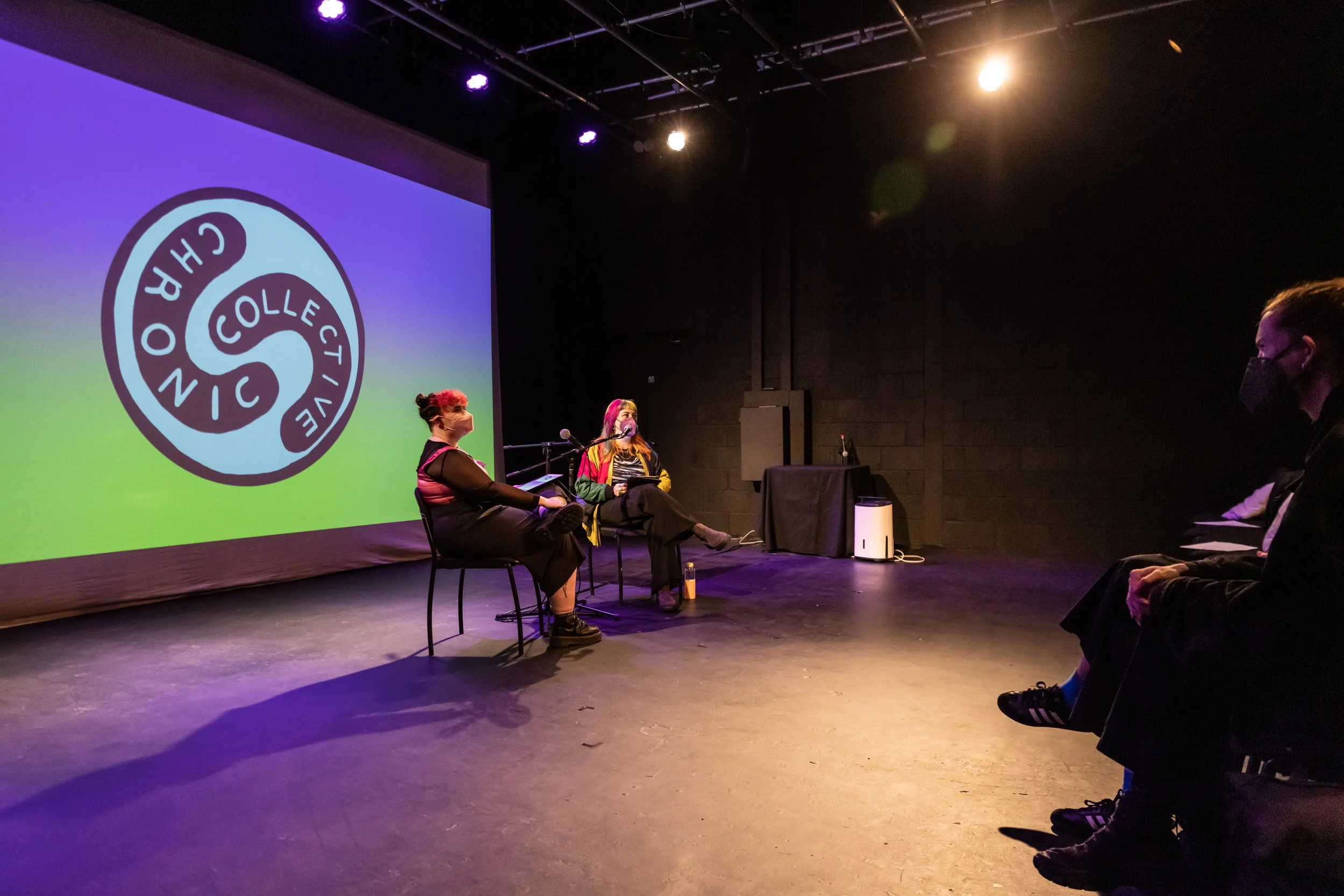 Two people are seated on stage in front of a large screen displaying a logo for 'Chronic Collective.' They are engaging in a discussion in a dimly lit room with an audience.