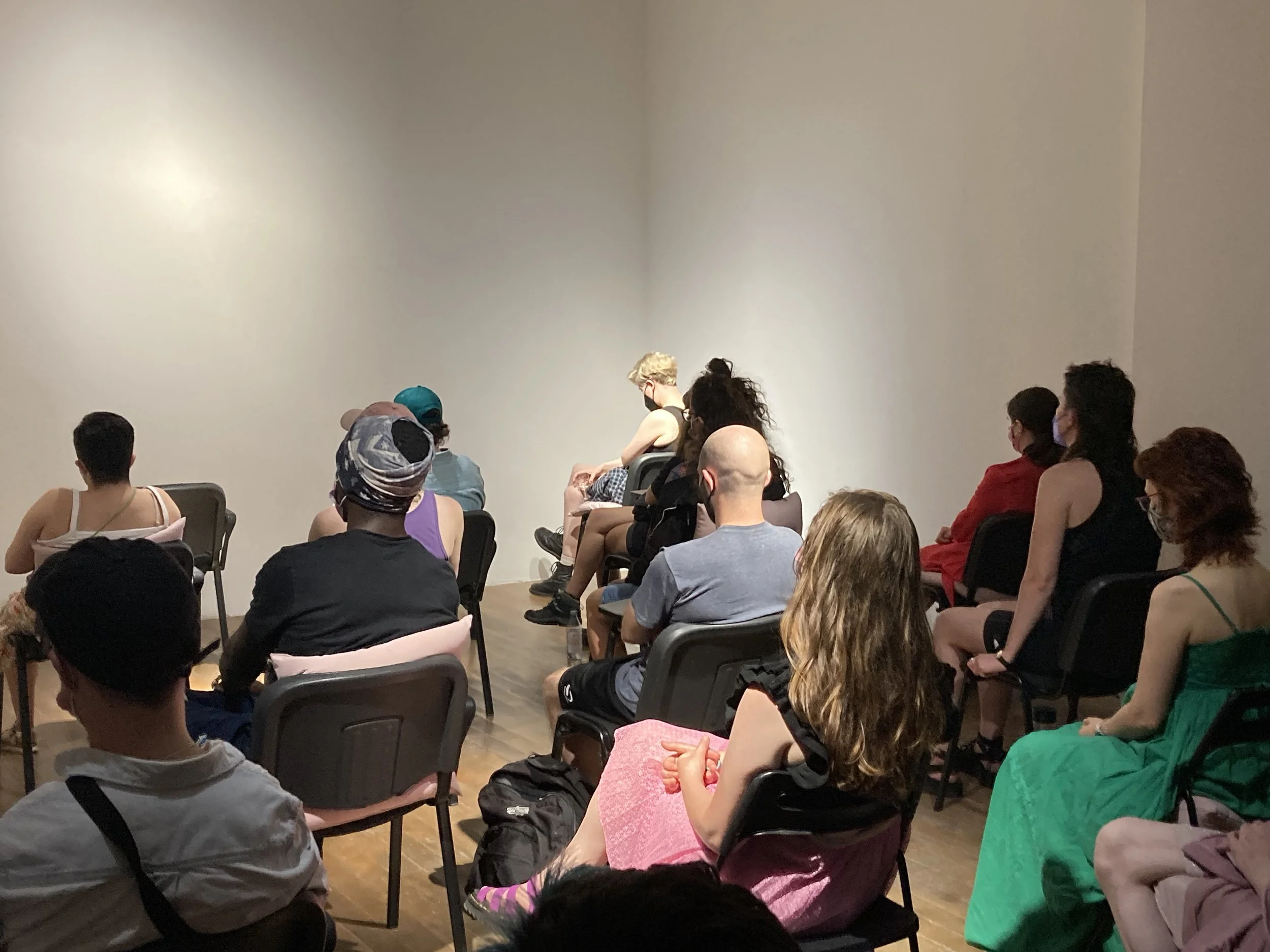 A group of people sitting on chairs facing a blank wall in a room, some wearing masks, attentively listening to a presentation or speaker.