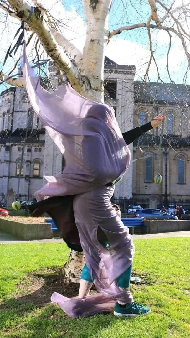 A person performing in a park with a tree, wearing a flowing purple material and teal sneakers, with a building and parked cars in the background.