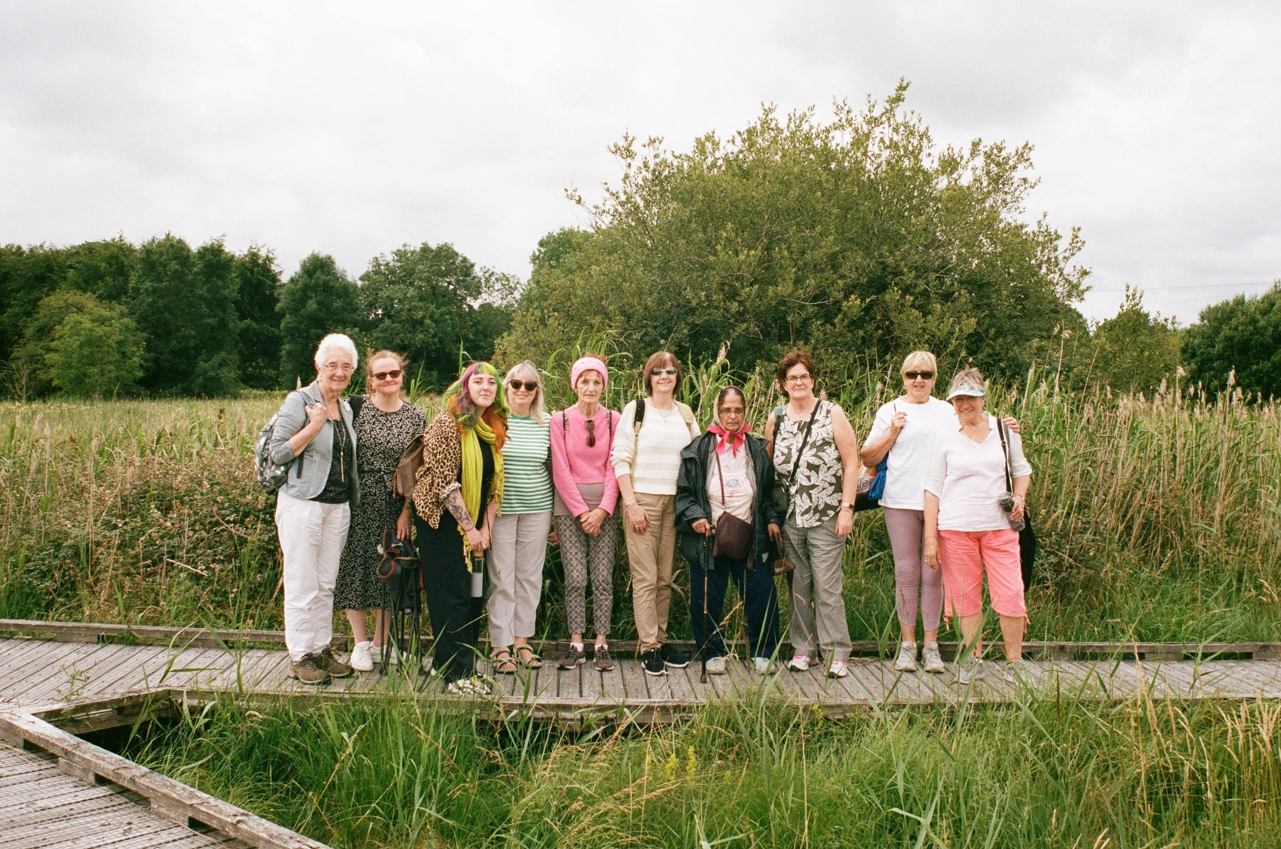 A group of twelve people standing together outdoors in a natural grassy field with trees and cloudy sky in the background.