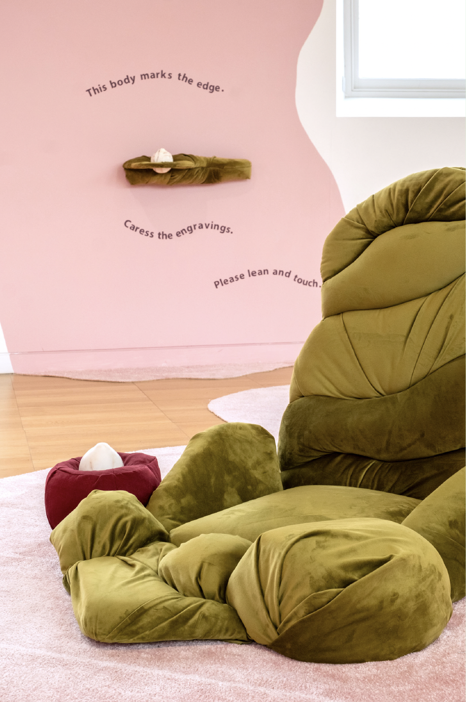 A cozy corner with a green velvet armchair and matching ottoman on a pink rug, next to a pink wall with wall art displaying text about touching and caressing engravings, and a shelf above with a small sculpture. A white window lets in light.