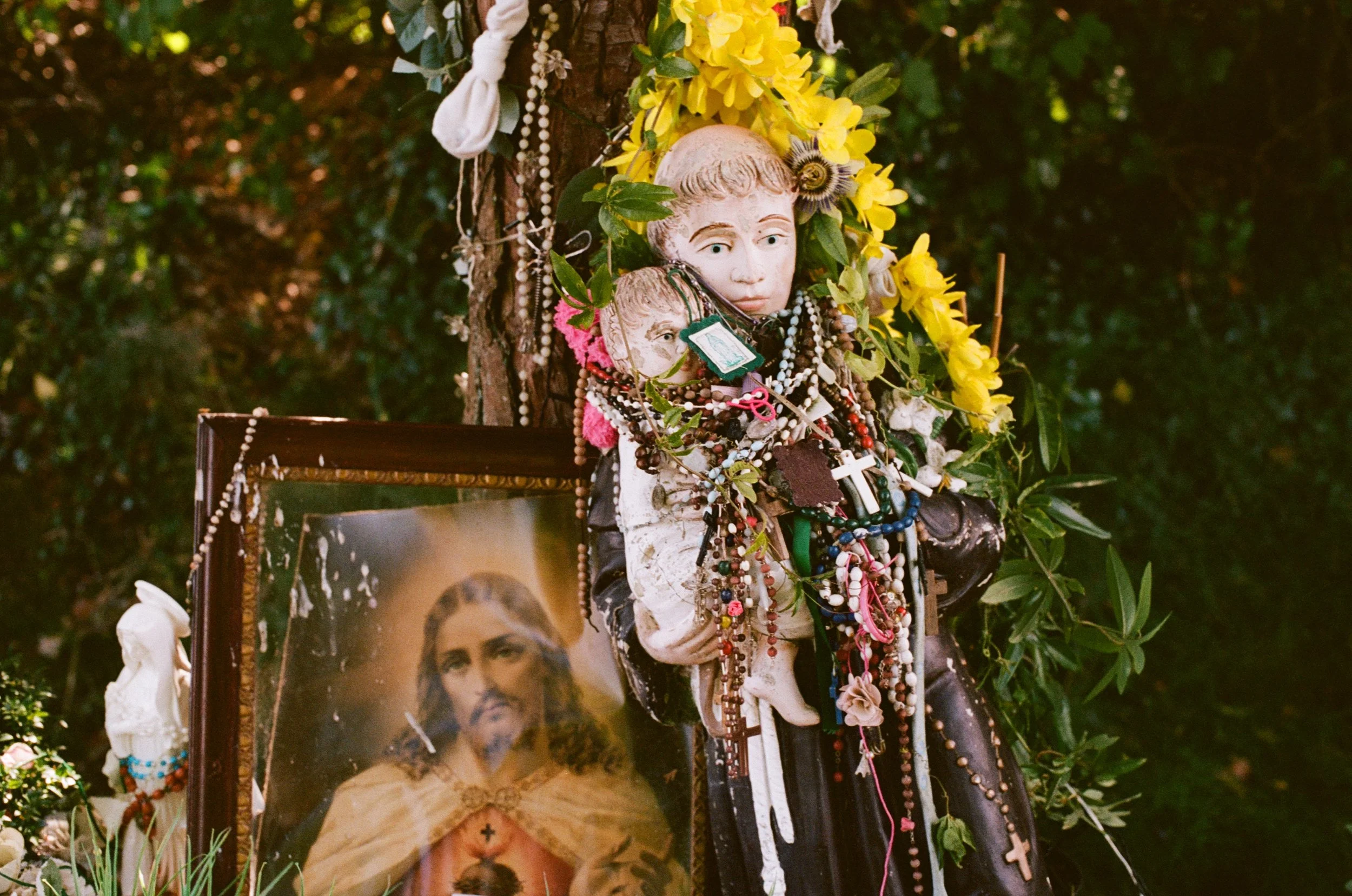 Religious shrine with a stone statue of a child decorated with rosaries, flowers, and jewelry, and a portrait of Jesus in the background.