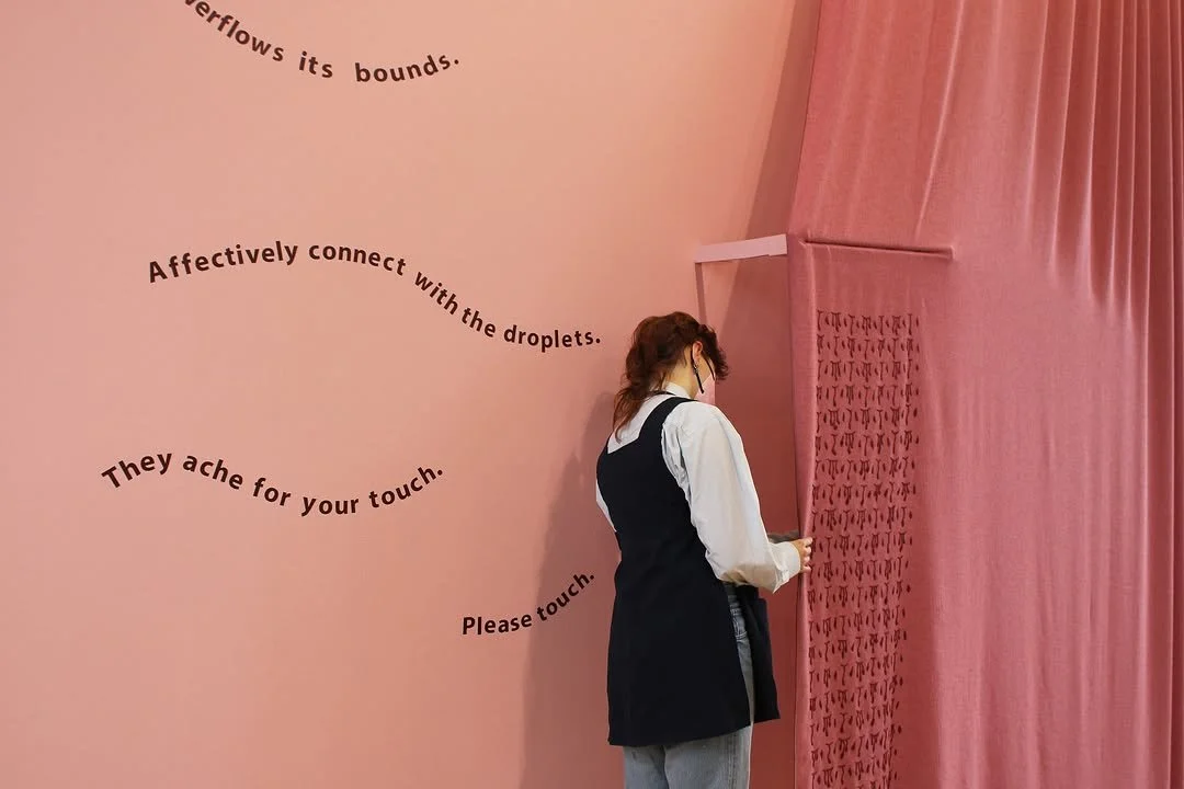 A person in a black vest and gray pants standing in front of a pink wall with purple curved text and a partially open pink curtain.