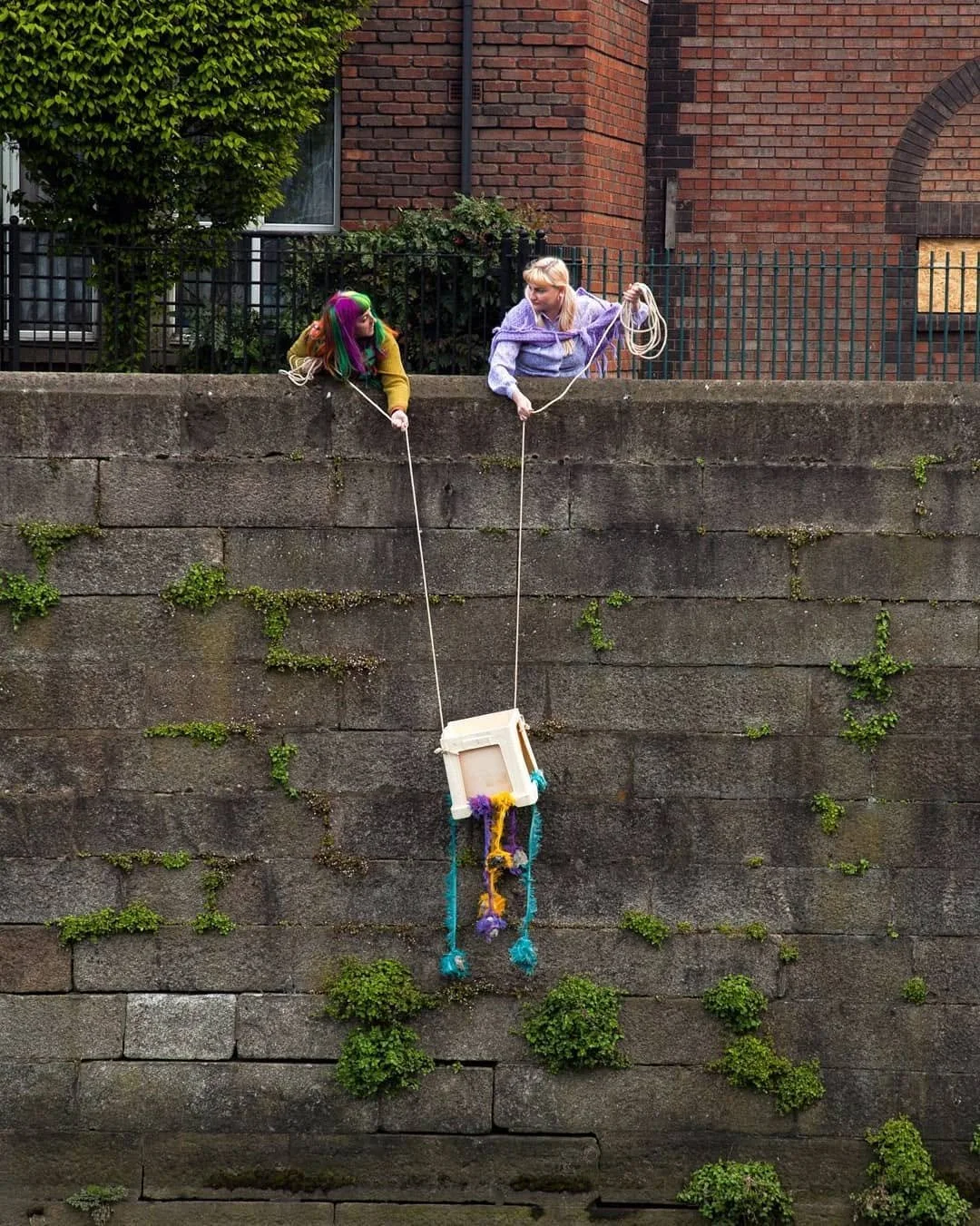 Two people with colorful hair leaning over a concrete wall, holding a rope that is attached to a small homemade swing decorated with yarn and fabric hanging down the side of the wall into the river.