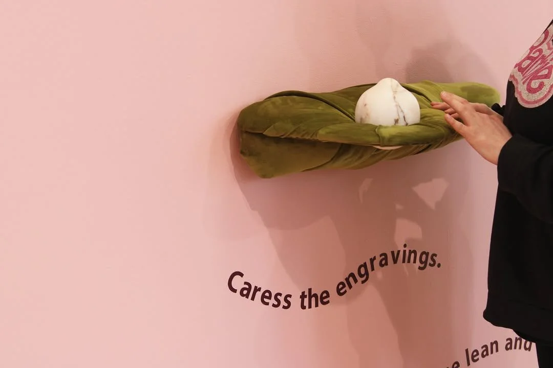 A person touching a marble sculpture resting on a green soft shelf display, with the phrase "Caress the engravings" on a pink wall.