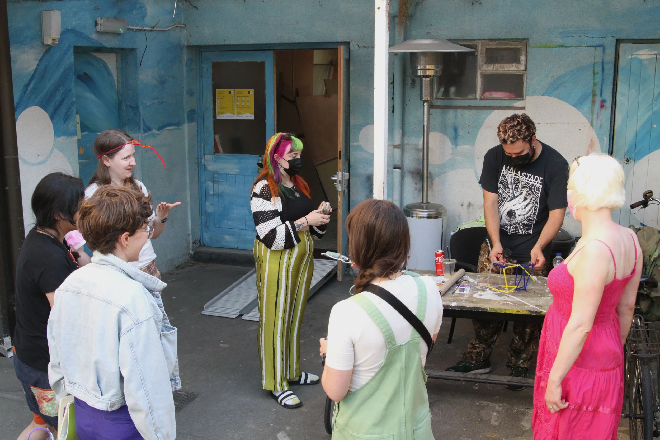 A group of people gathered outdoors around a table and a person performing sitting on a table. Some people are wearing masks, and one person has colourful hair.