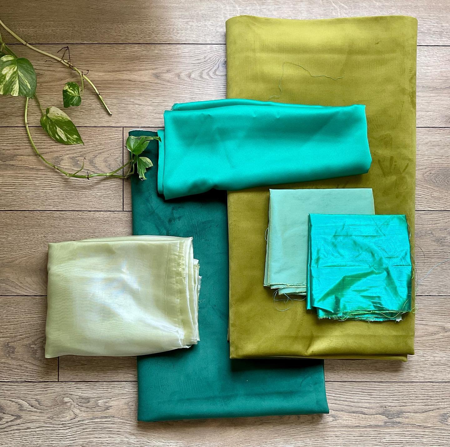 Various pieces of fabric in shades of yellow, green, and blue are neatly arranged on a wooden surface, with a small green leafy plant in the top left corner.