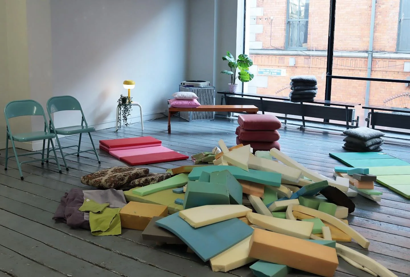 An art studio with foam pieces scattered on the wooden floor, chairs stacked against the wall, a small table with a yellow lamp, green plant near a large window, and folded fabrics on a bench.