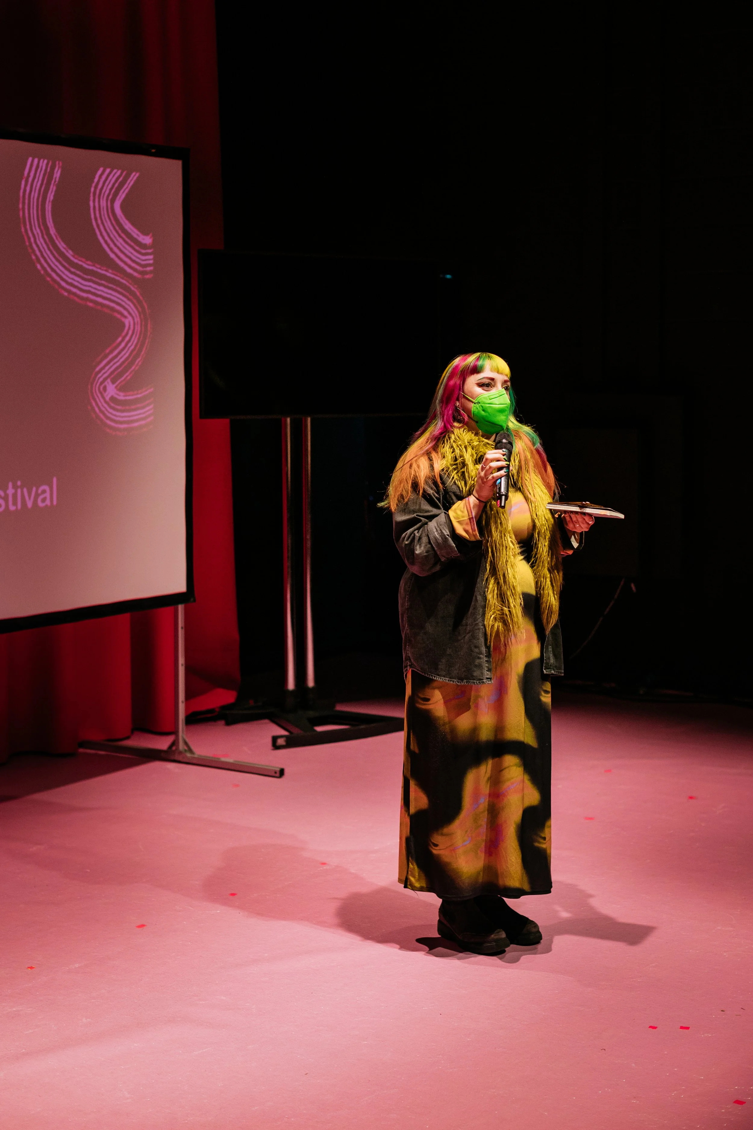 A person wearing a colorful dress, a green mask, and a black jacket, holding a microphone and some papers, standing on a stage with a pink floor and a large screen displaying a pink and purple logo behind them.