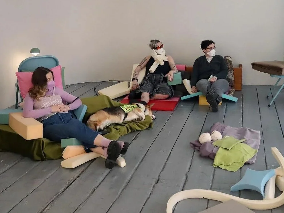 Three people and a dog resting in an art workshop made of colorful foam blocks and cushions, with scattered fabric and objects on a wooden floor.