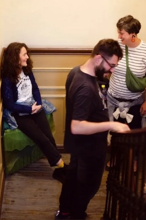 Three people in a room. One person is sitting on a green bench, smiling, and wearing a white shirt with a design, a navy jacket, and black pants. Two other people are standing; one with glasses and a beard, looking down at a device, and the other with short hair, wearing a striped shirt, orange earrings, and a green bag, smiling while looking at the person with glasses.