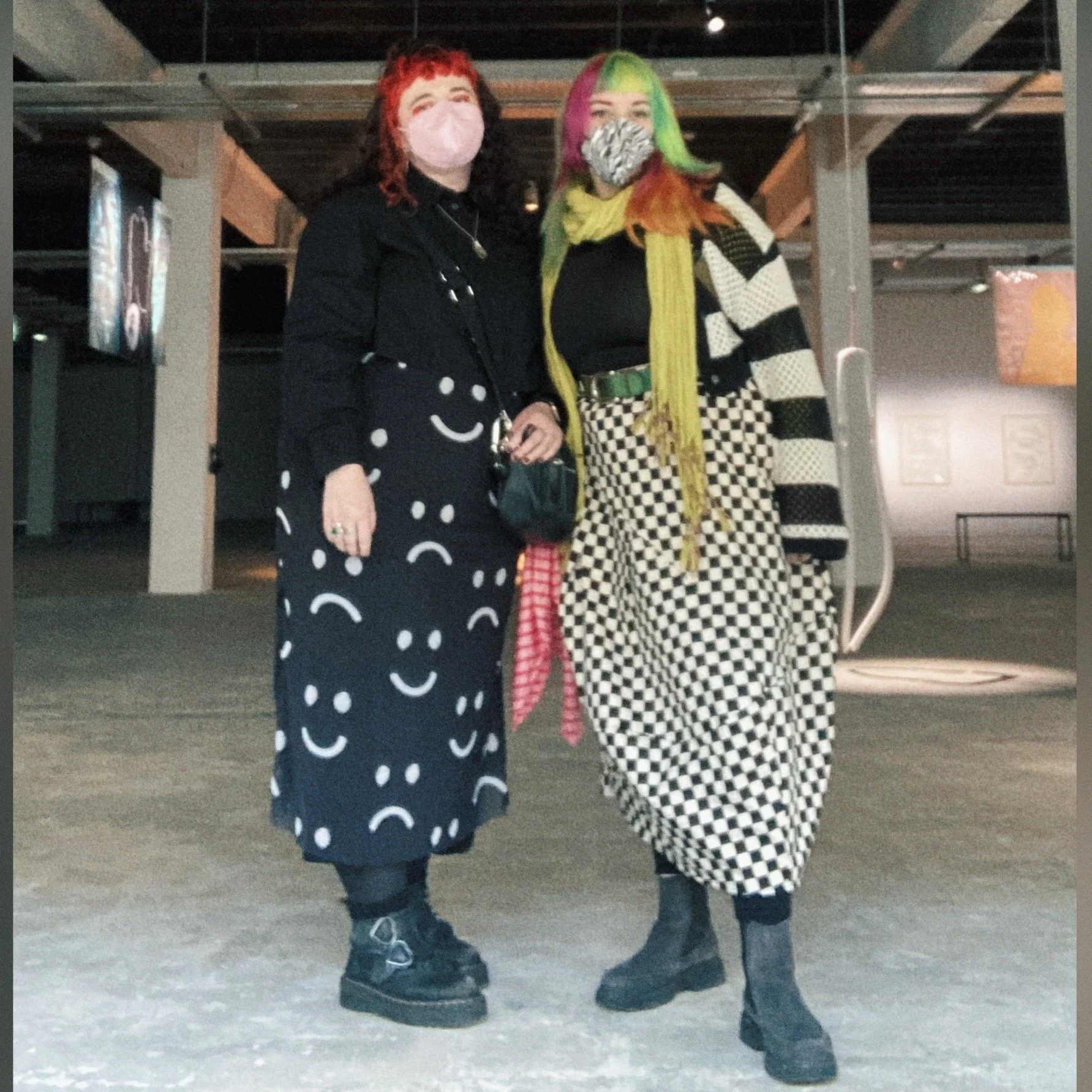 Two people standing side by side, wearing colourful attire and masks, indoors with a modern industrial ceiling and some wall art visible in the background.
