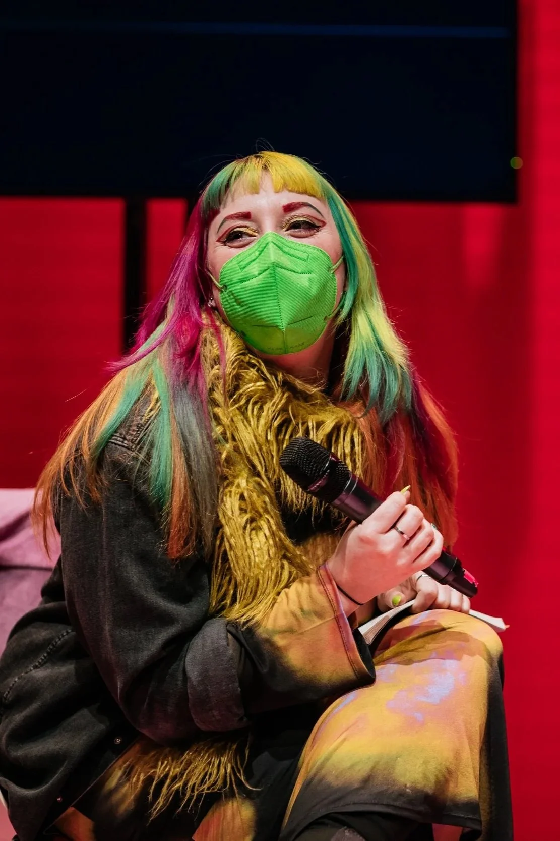 A person with multi-colored hair wearing a green face mask, sitting on a stage, holding a microphone and smiling.