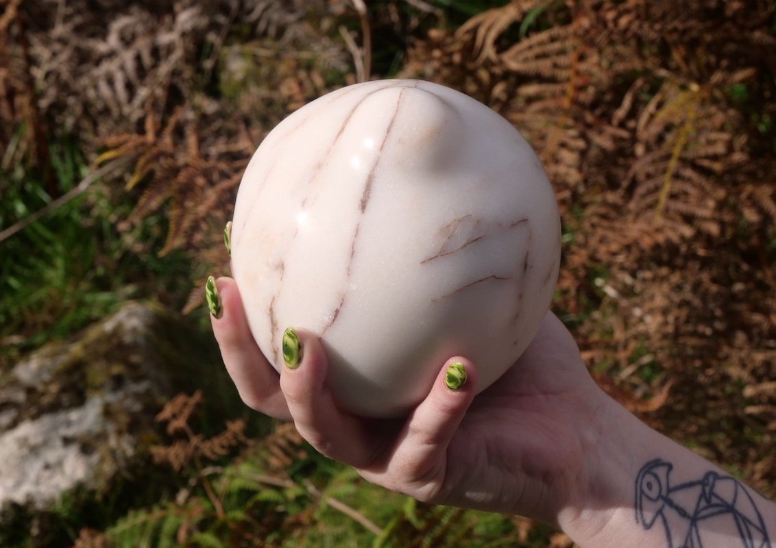 A hand with green and black painted nails holding a large white marble sculpture with brown veining outdoors among brown and green foliage.