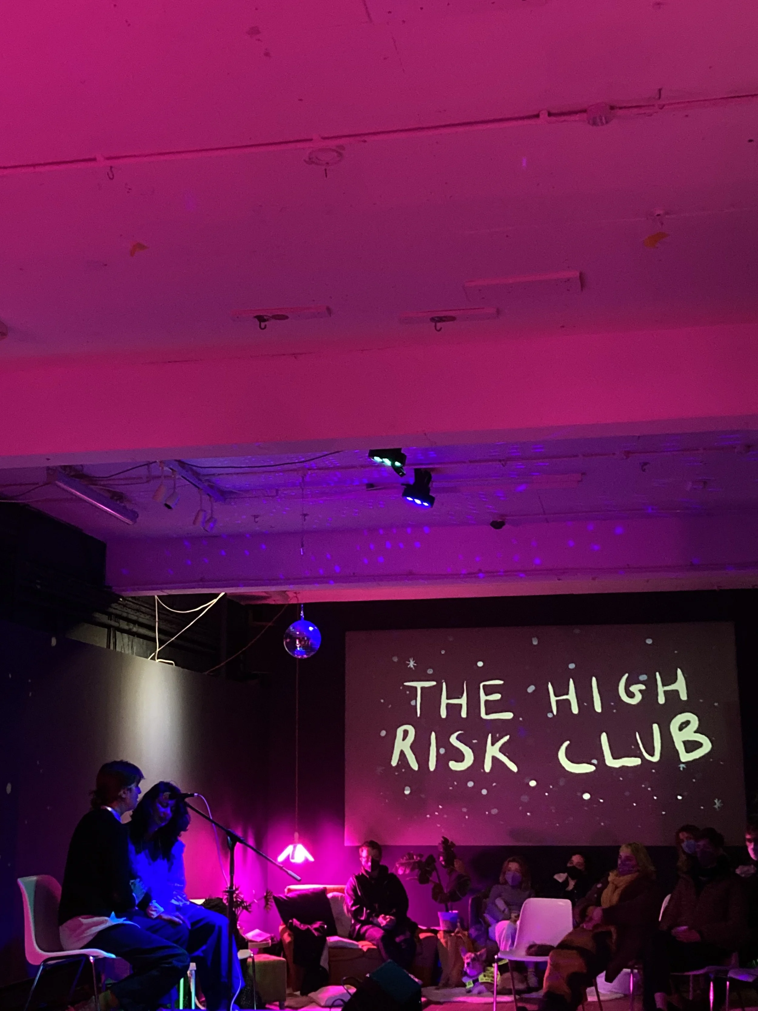 People sitting and standing in a dimly lit room with purple and pink lighting, some with microphones, and a large banner that reads 'THE HIGH RISK CLUB' in the background.