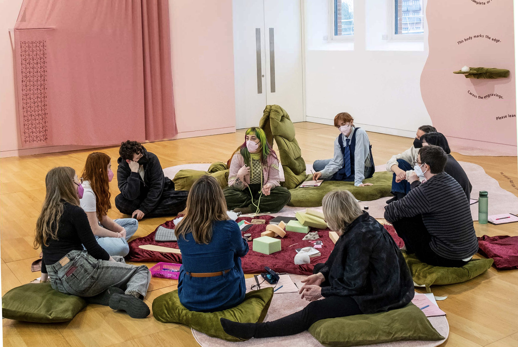 Group of people sitting on cushions in a circle in an art gallery or museum, engaging in a discussion or workshop, with some wearing masks, and various objects and notebooks on the floor around them.