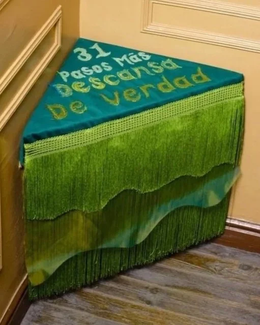 A decorative piece in the shape of a table with a colorful cloth covering it. The cloth has a green background with a gradient and fringe at the bottom. The top of the table has text written in yellow and white that says '31 Pasos Más Desconcertar De Verdad'.