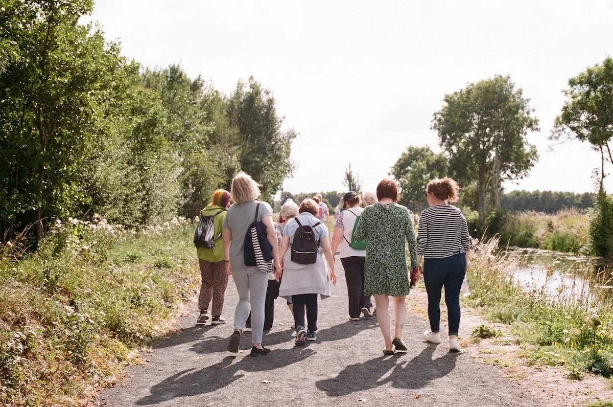 Group of people walking along a nature trail beside a river with trees and grass on a sunny day.