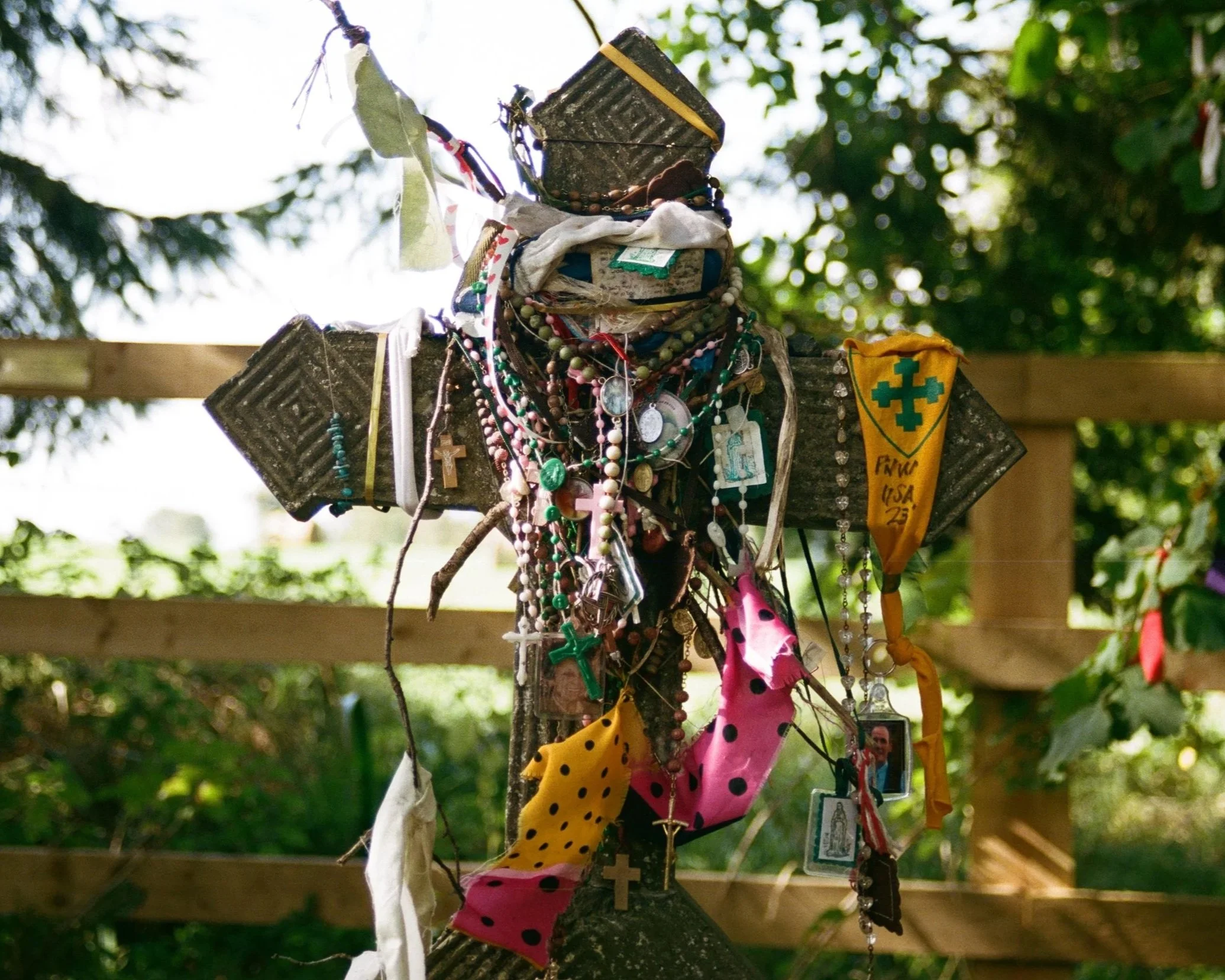 A wooden cross decorated with various necklaces, beads, scarves, and small items, adorned with a yellow cloth featuring a green cross, set outdoors against a backdrop of green foliage and sunlight.