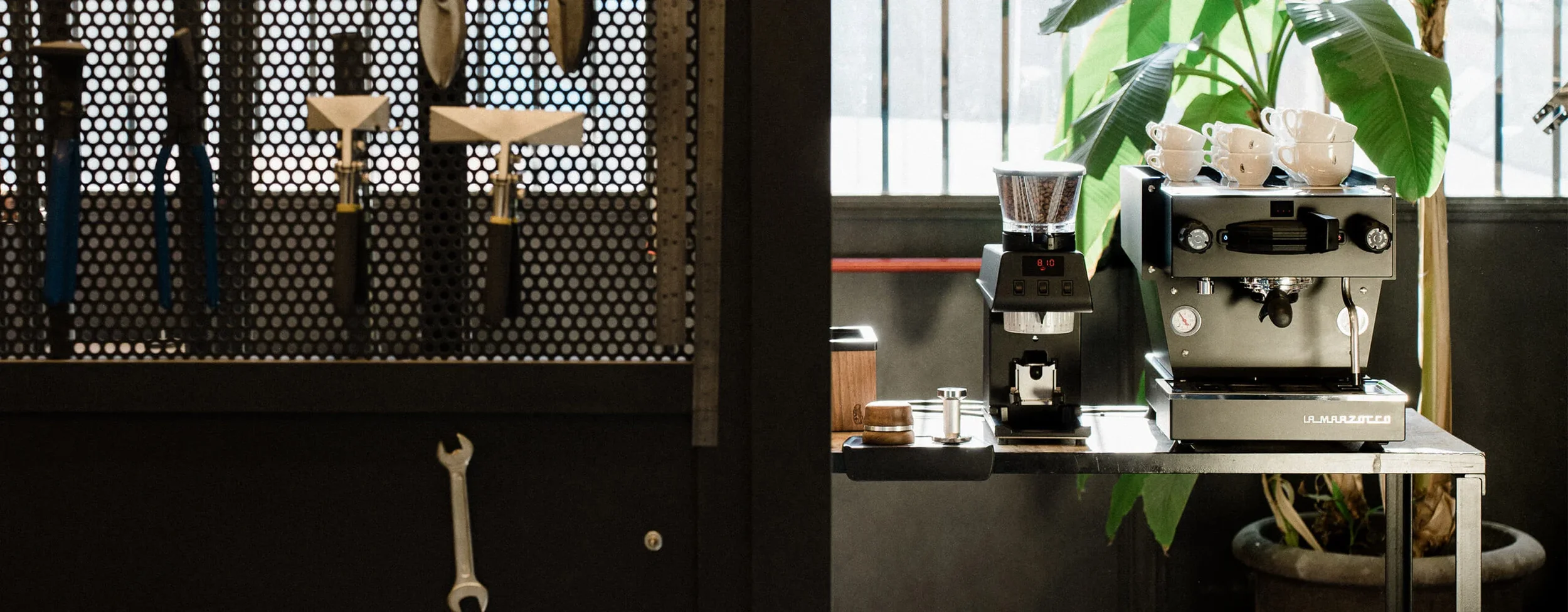 Ein Kaffeevollautomat mit weißen Kaffeetassen auf der Oberseite, eine Kaffeemühle, einige Werkzeuge und Pflanzen im Hintergrund, in einer modernen Kaffee- oder Espressobar.