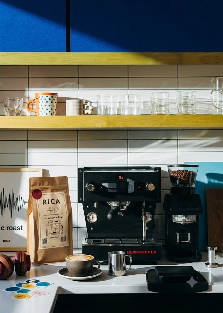 Kaffeebereiter und Zubehör auf einer weißen Theke, darunter eine Tasse Cappuccino, eine Metallmilchkanne, eine Packung RICA Kaffee, eine Kaffeemaschine, eine Kapselmaschine und eine Brille mit Kopfhörern.