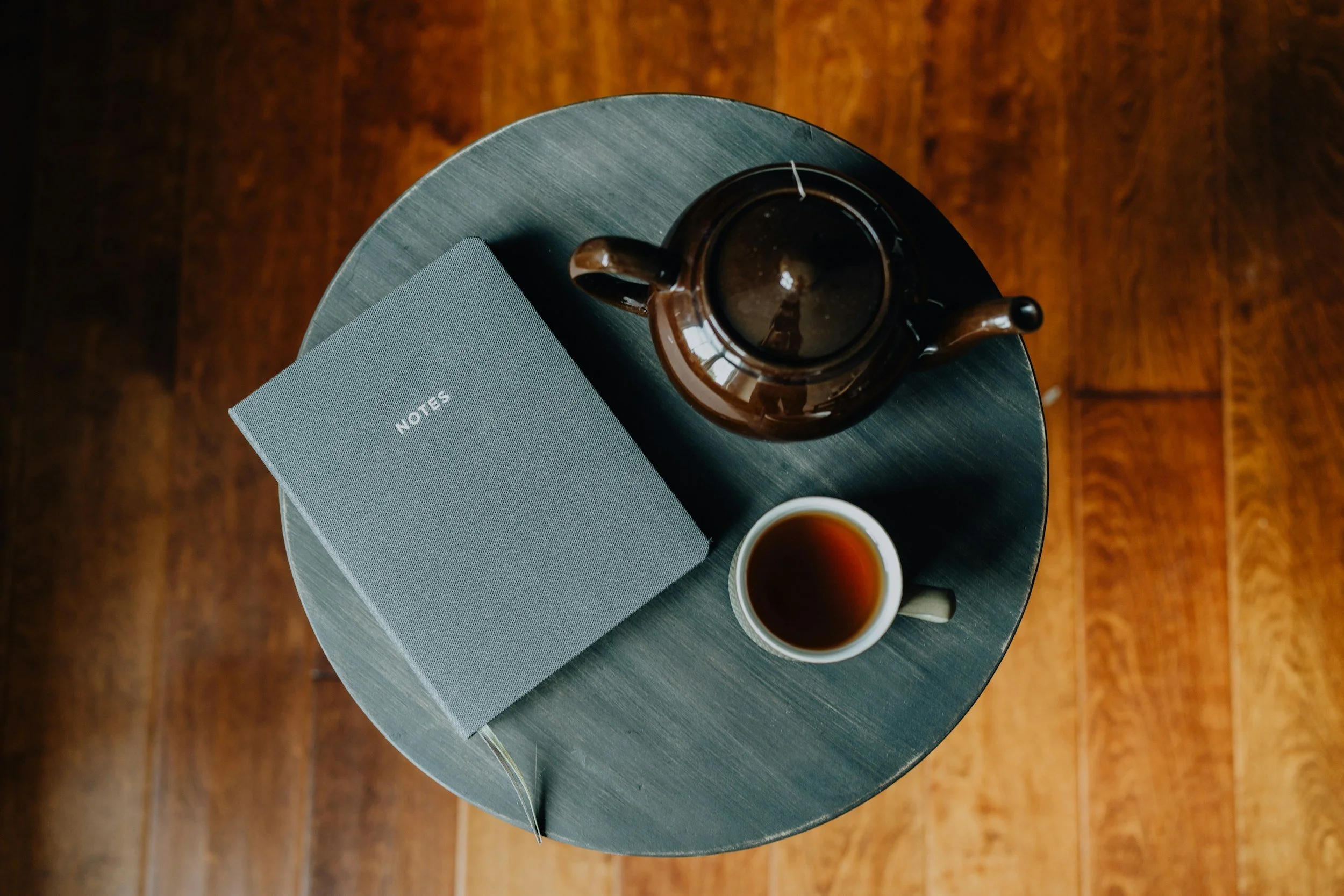 Top-down view of a round table with a dark teapot, a notebook labeled 'NOTES', and a cup of tea on a gray surface, with a wooden floor in the background.
