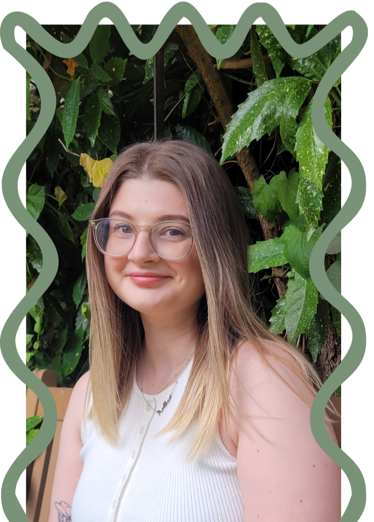 A young woman with long, blonde hair and glasses smiling outdoors in front of lush green leafy plants.