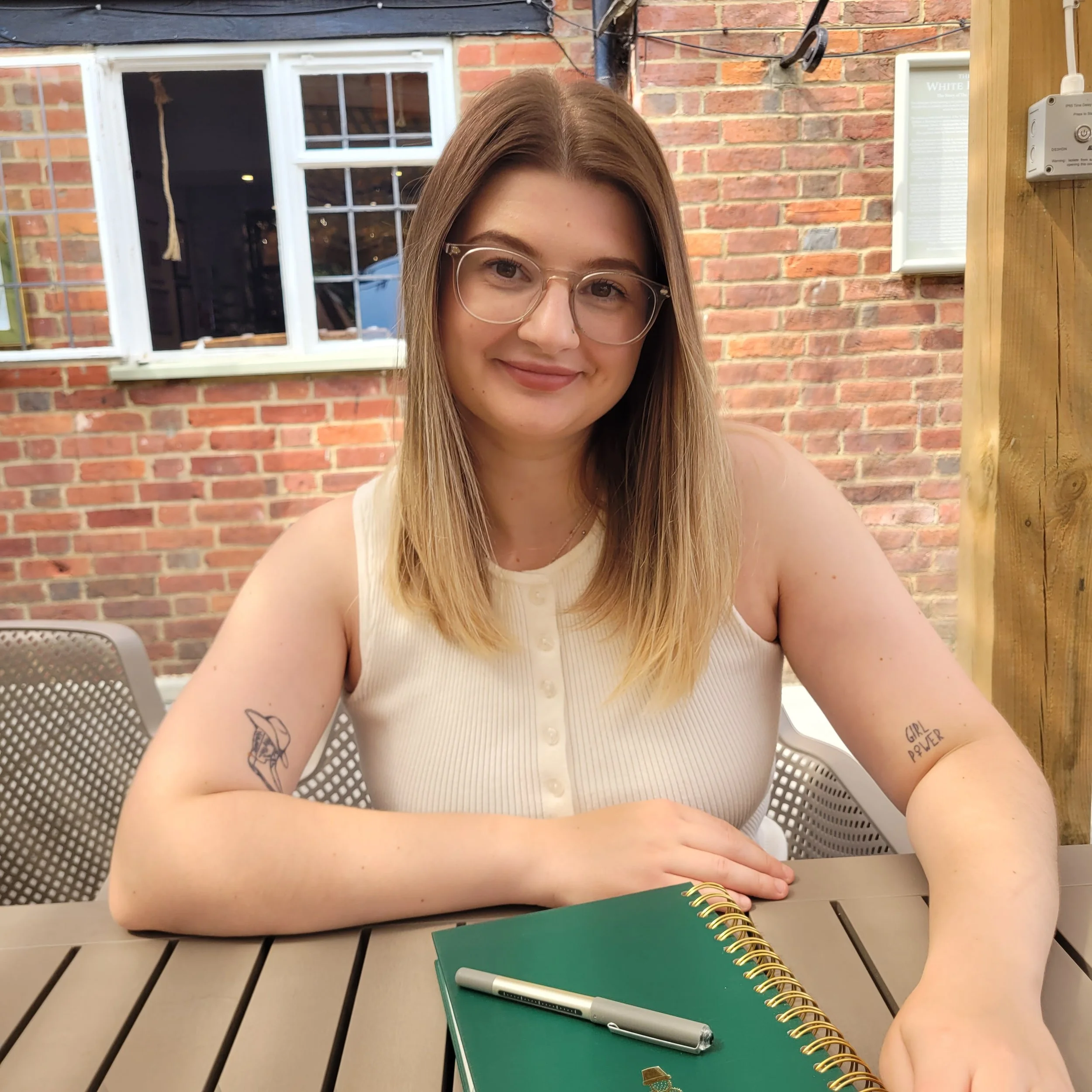 A young woman with long blonde hair, glasses, and a nose piercing smiling at the camera sitting at a table outside with a brick wall and a window behind her. She has tattoos on her arms, with one reading 'Girl Power'. On the table, there is a green notebook and a silver pen, with another pen near her hand.