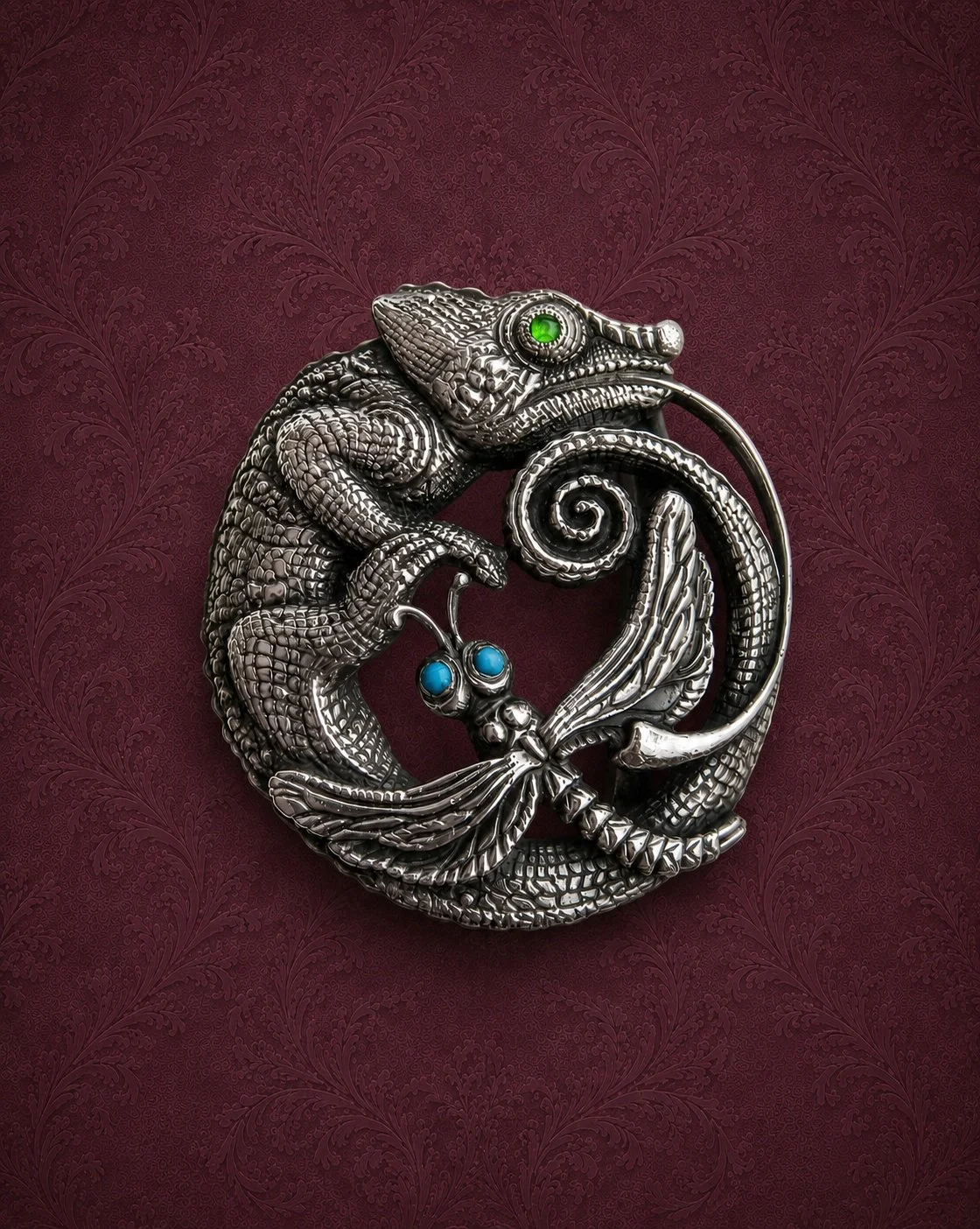 A silver dragon-shaped jewelry piece with green and blue gemstones, set against a red patterned background.