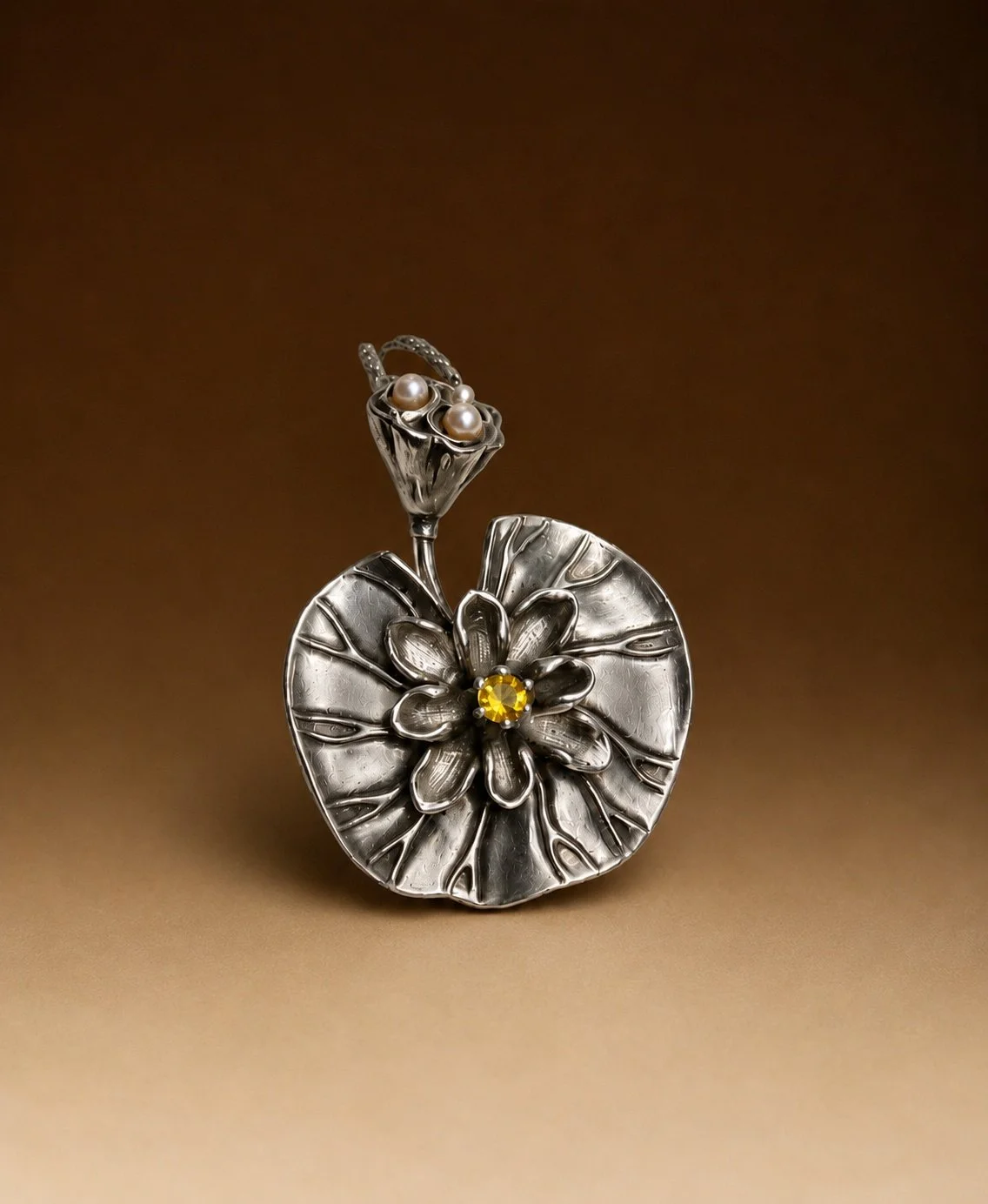 Silver decorative brooch with a floral and leaf design, featuring pearl and yellow gemstone embellishments against a brown background.