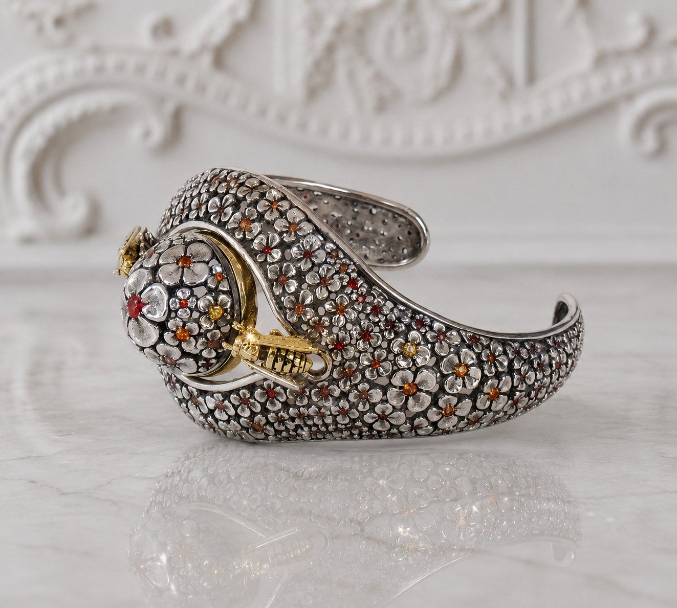 A silver and gold floral-embellished bracelet with a floral clasp, decorated with small gemstones, is on a reflective surface with a decorative white background.