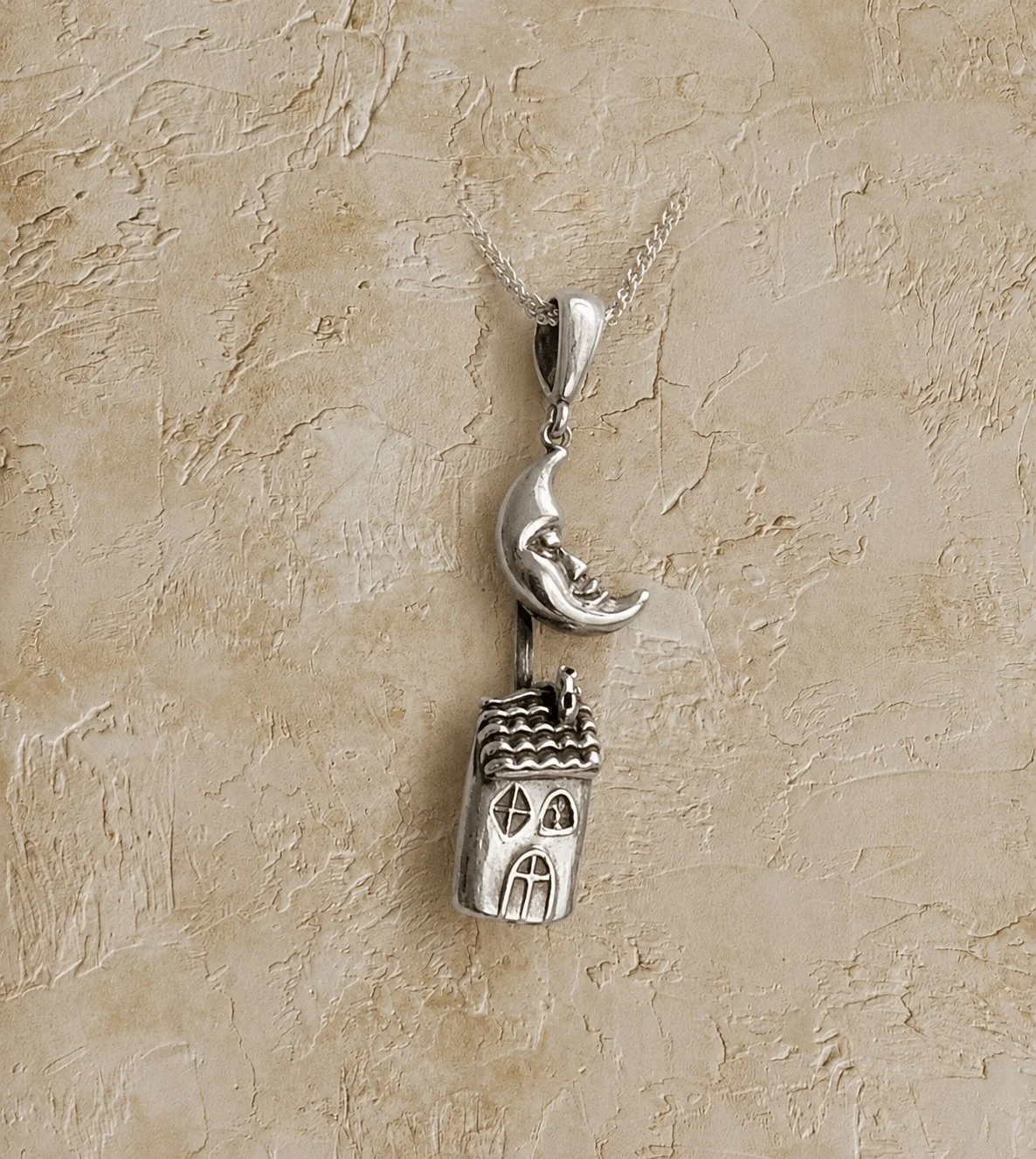 Silver necklace with a pendant featuring a crescent moon with a face and a small house with windows, on a textured beige background.