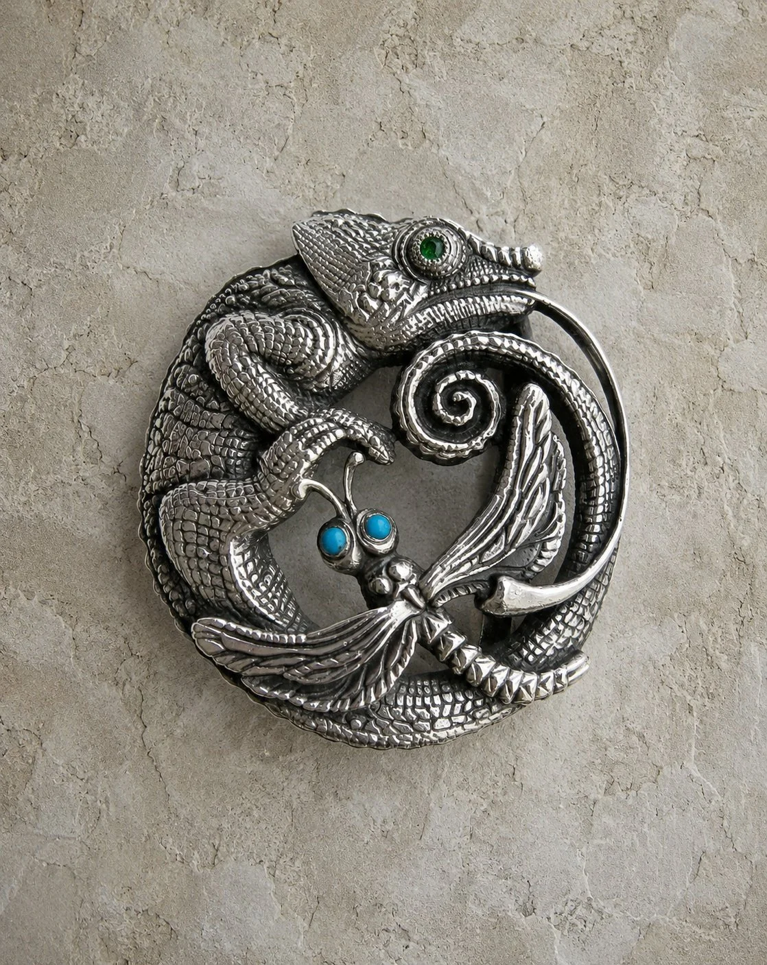 A silver jewelry piece shaped like a chameleon with a curled tail and a butterfly with blue gemstone eyes, both intricately designed with textured scales and wings.
