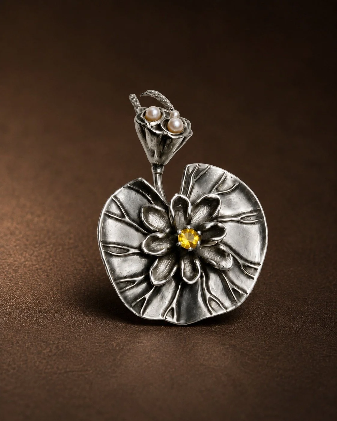 A silver jewelry piece resembling a flower with a large circular base, a central flower design with a yellow gemstone, and a stem ending in a leaf with pearls in a cup-shaped setting, displayed on a brown surface.