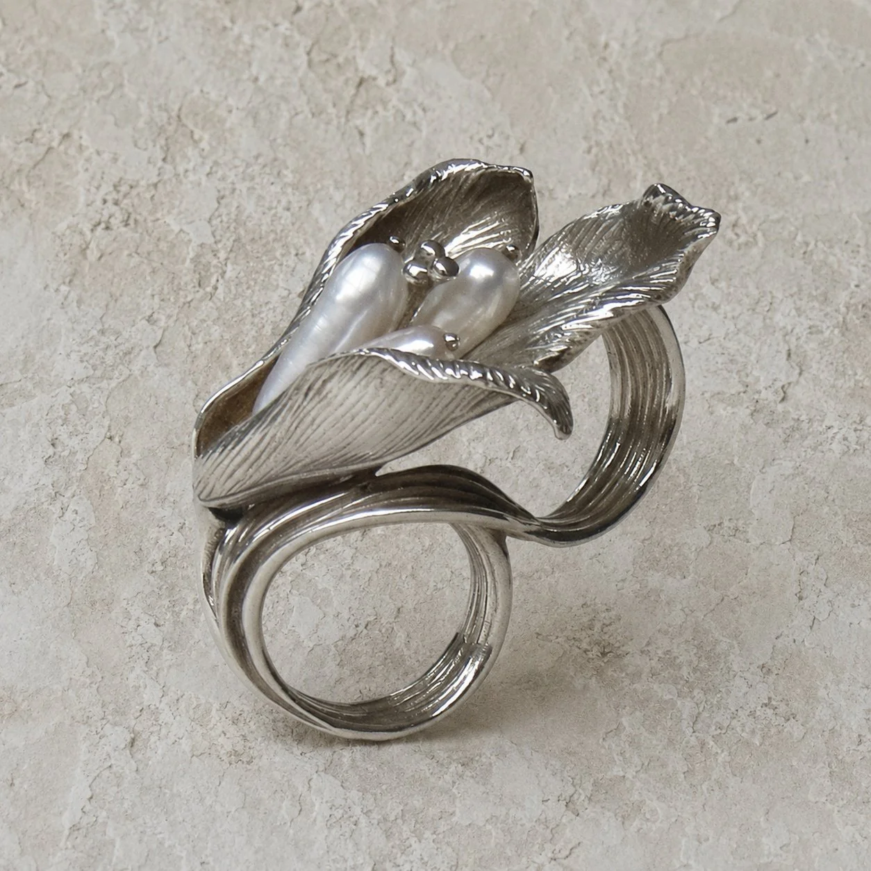 Silver ring shaped like a flower with metallic petals and pearl accents on a textured beige surface.