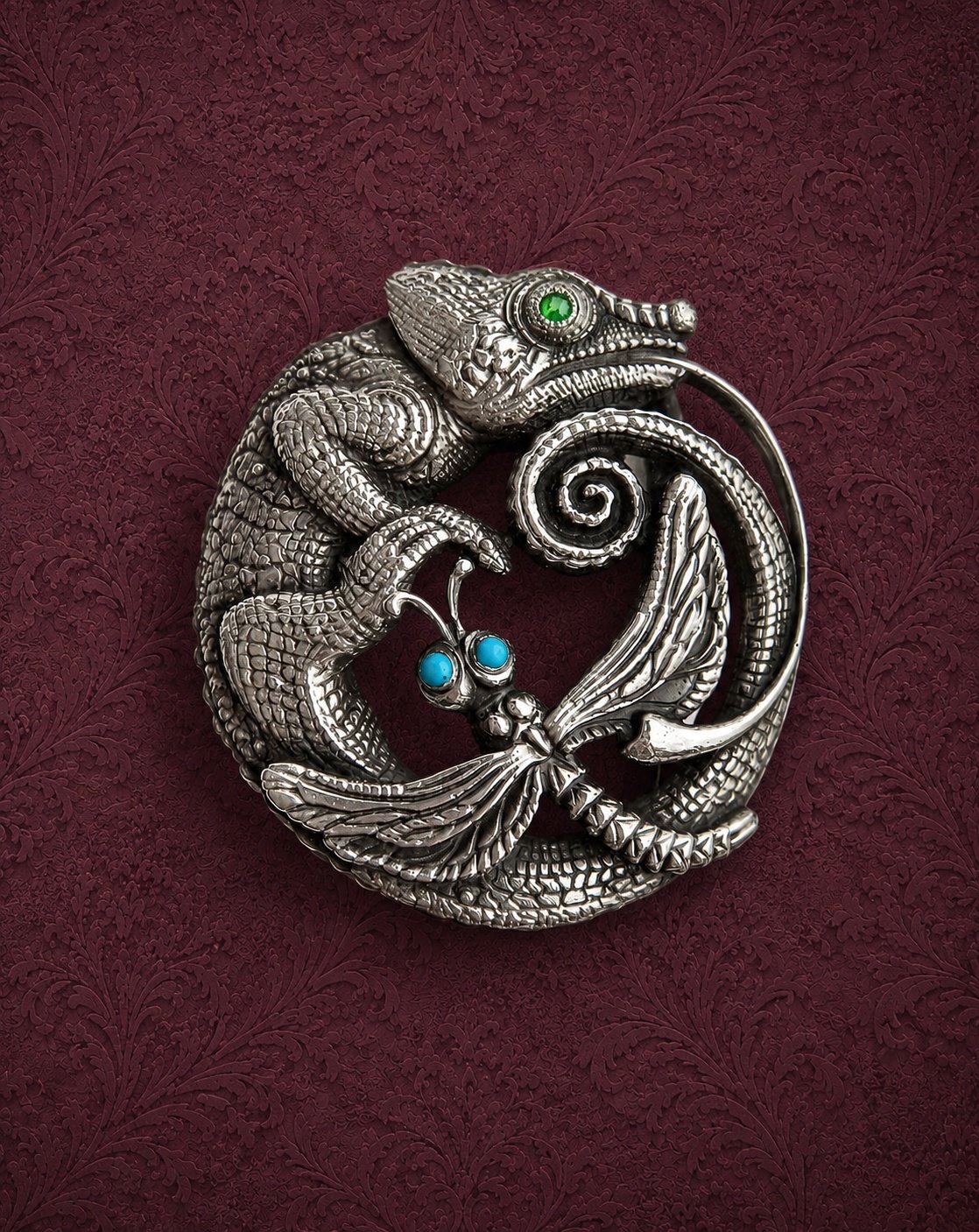 Silver brooch featuring a chameleon and a butterfly with colored gemstones for eyes, placed on a textured maroon background.