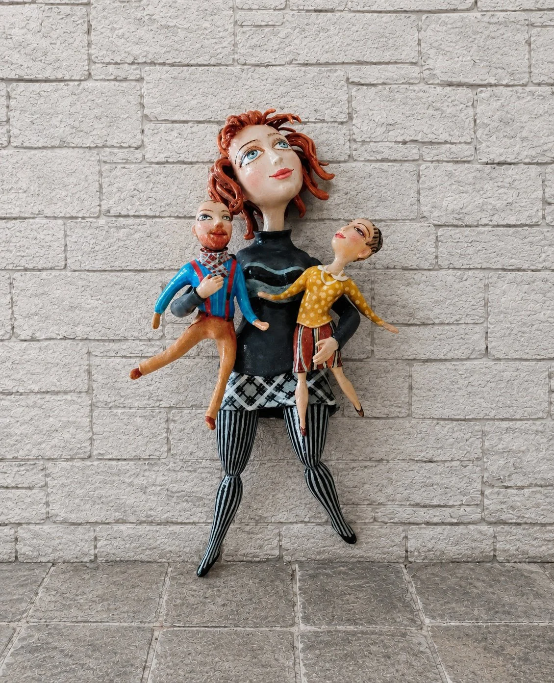 Colorful sculpture of a woman with two children, all with exaggerated features, standing against a light gray brick wall.