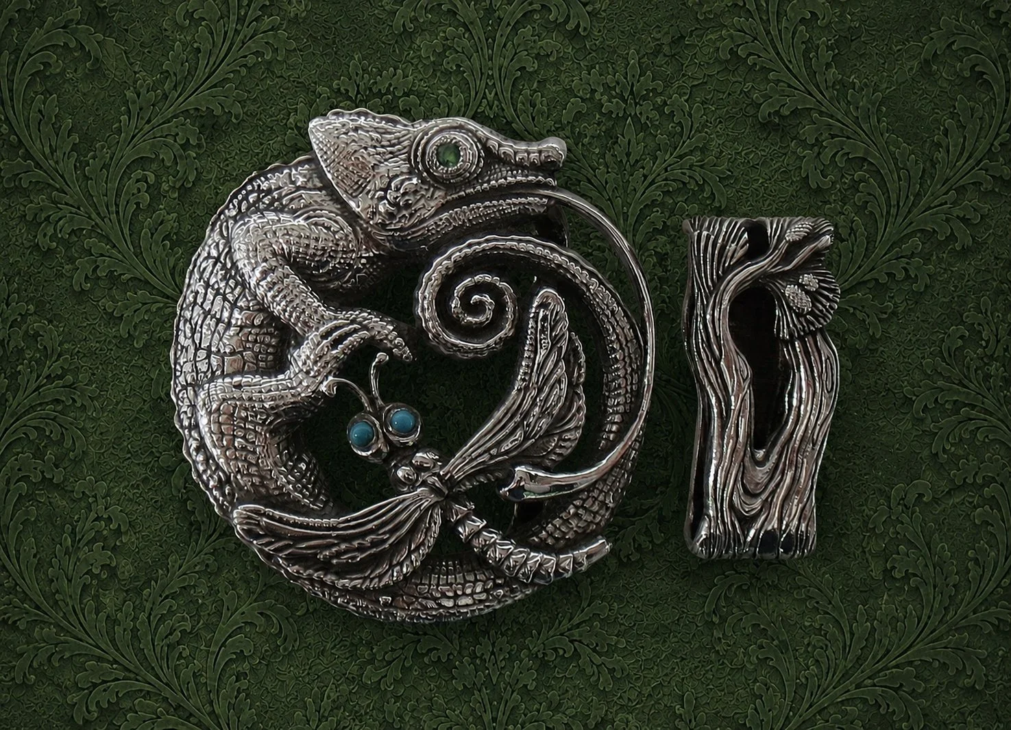 A silver chameleon-shaped brooch with teal eyes, holding a branch with berries, placed next to a wooden money clip, on a dark green textured background with a leafy pattern.